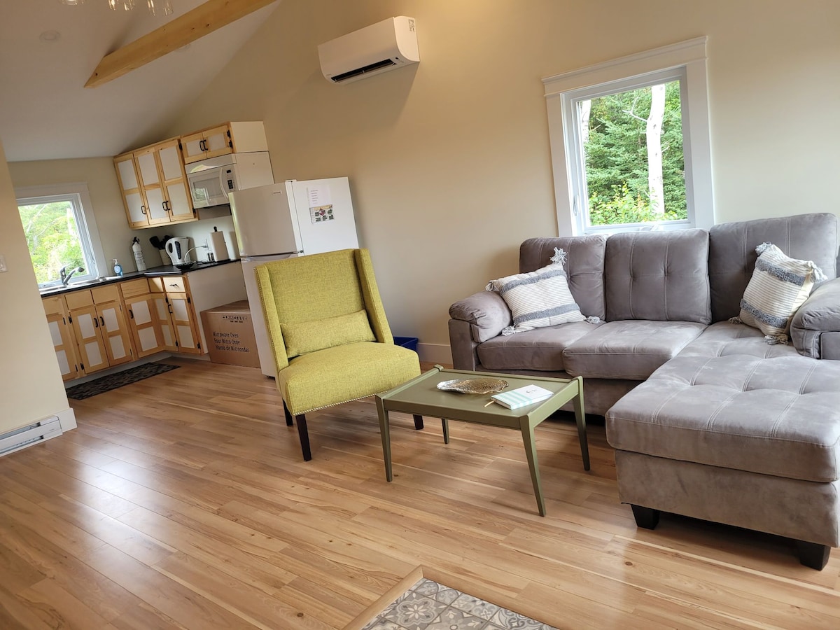 A cozy living area features a plush sectional sofa and a green accent chair, inviting relaxation. Natural wood flooring complements light-colored walls. A small coffee table sits in front of the sofa, and the kitchen area is visible in the background with wooden cabinets and modern appliances.