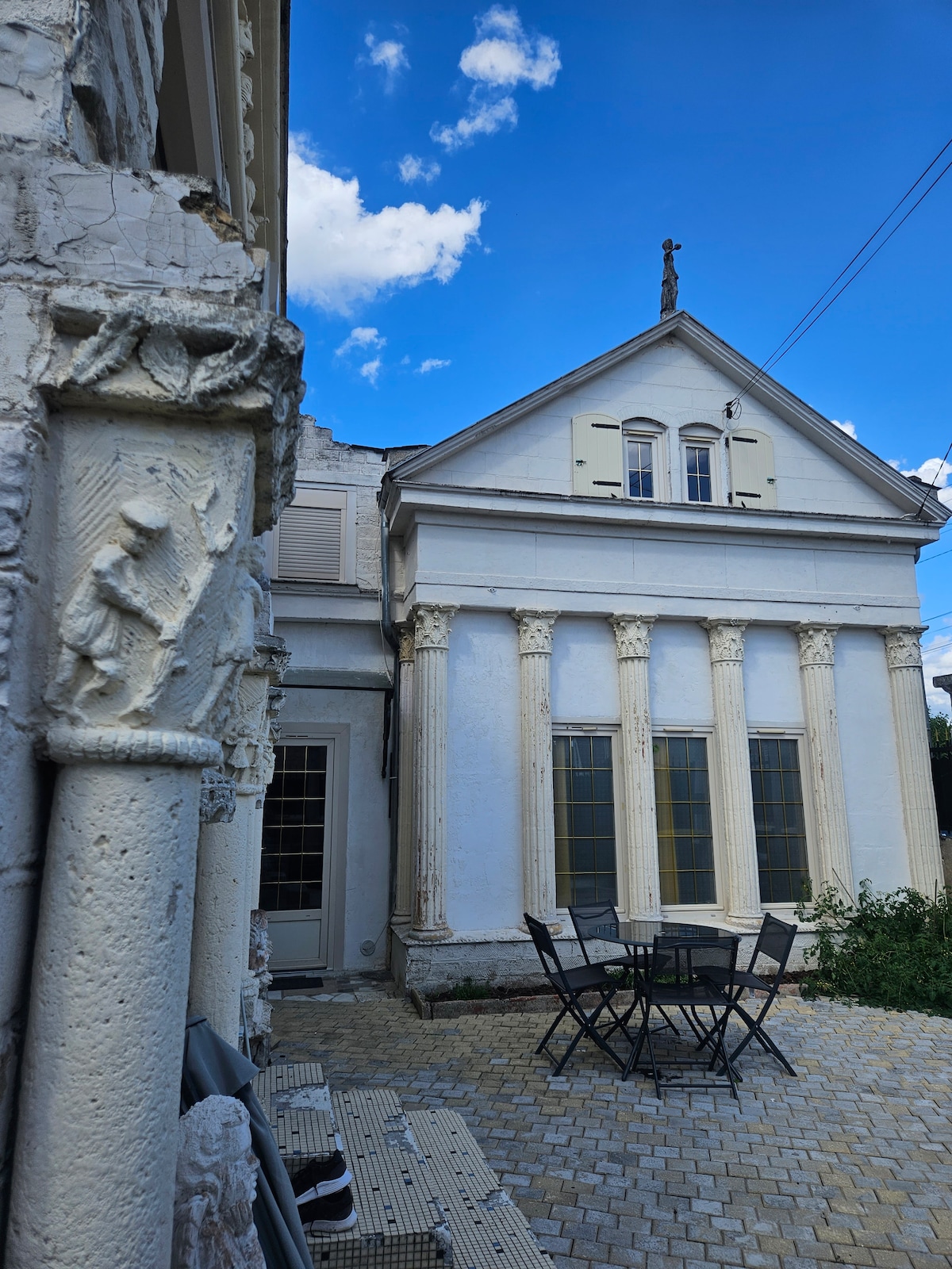 The image displays the exterior of a unique duplex with architectural features, including columns and intricate carvings. A small patio with a round dining table and chairs is visible, surrounded by greenery. The sky above is bright and partly cloudy.