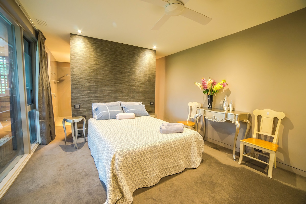 A spacious bedroom is highlighted by neutral-colored walls and a textured feature wall. A queen size bed is adorned with soft linens, accompanied by a nightstand and a table set with a vase of flowers. Natural light filters through large glass doors leading to the outdoors.
