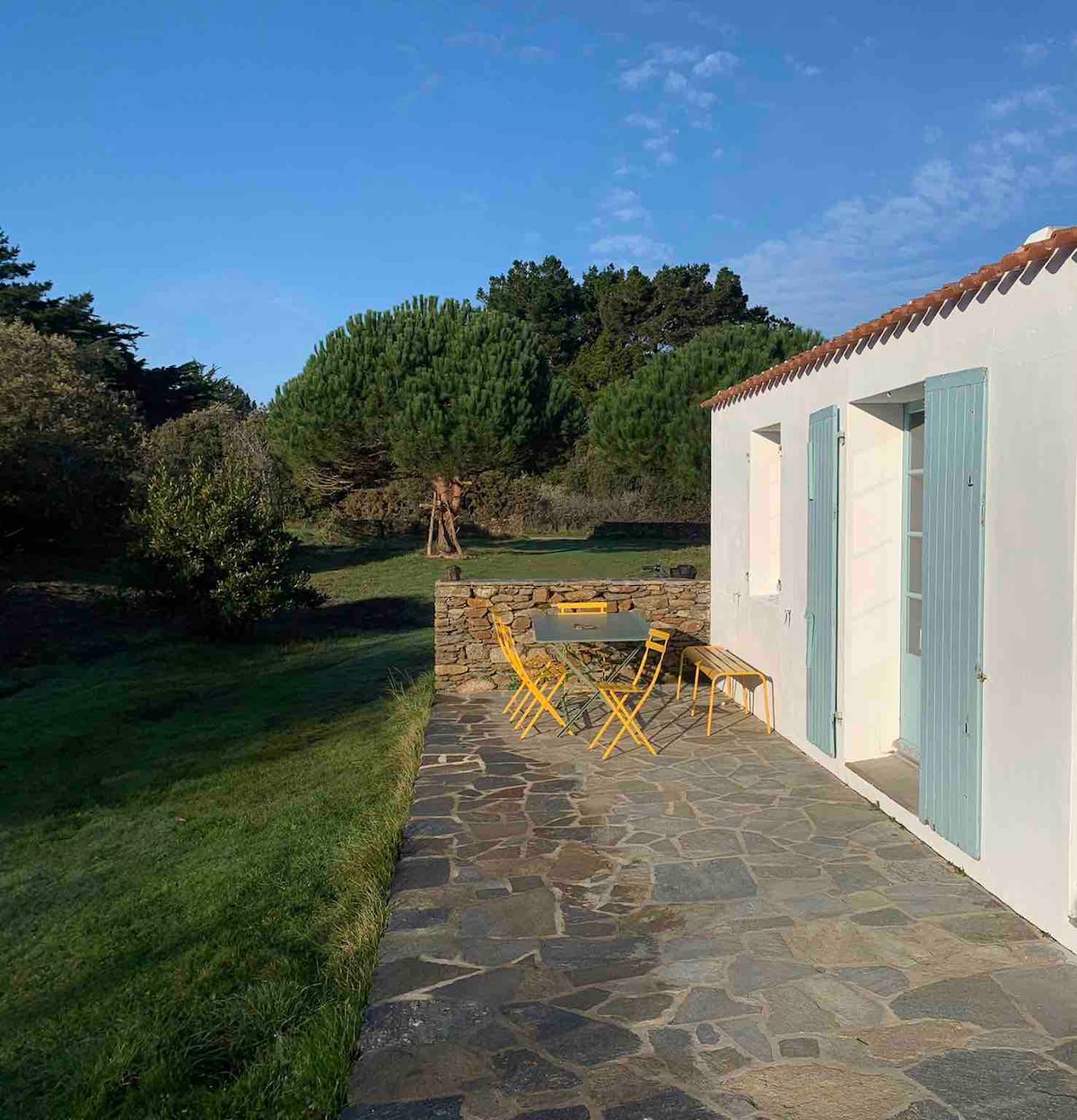 An outdoor terrace features a stone floor and a wooden table surrounded by yellow chairs, overlooking a green garden. The light-colored walls of the house are complemented by pastel shutters, while tall trees are visible in the background against a clear blue sky.