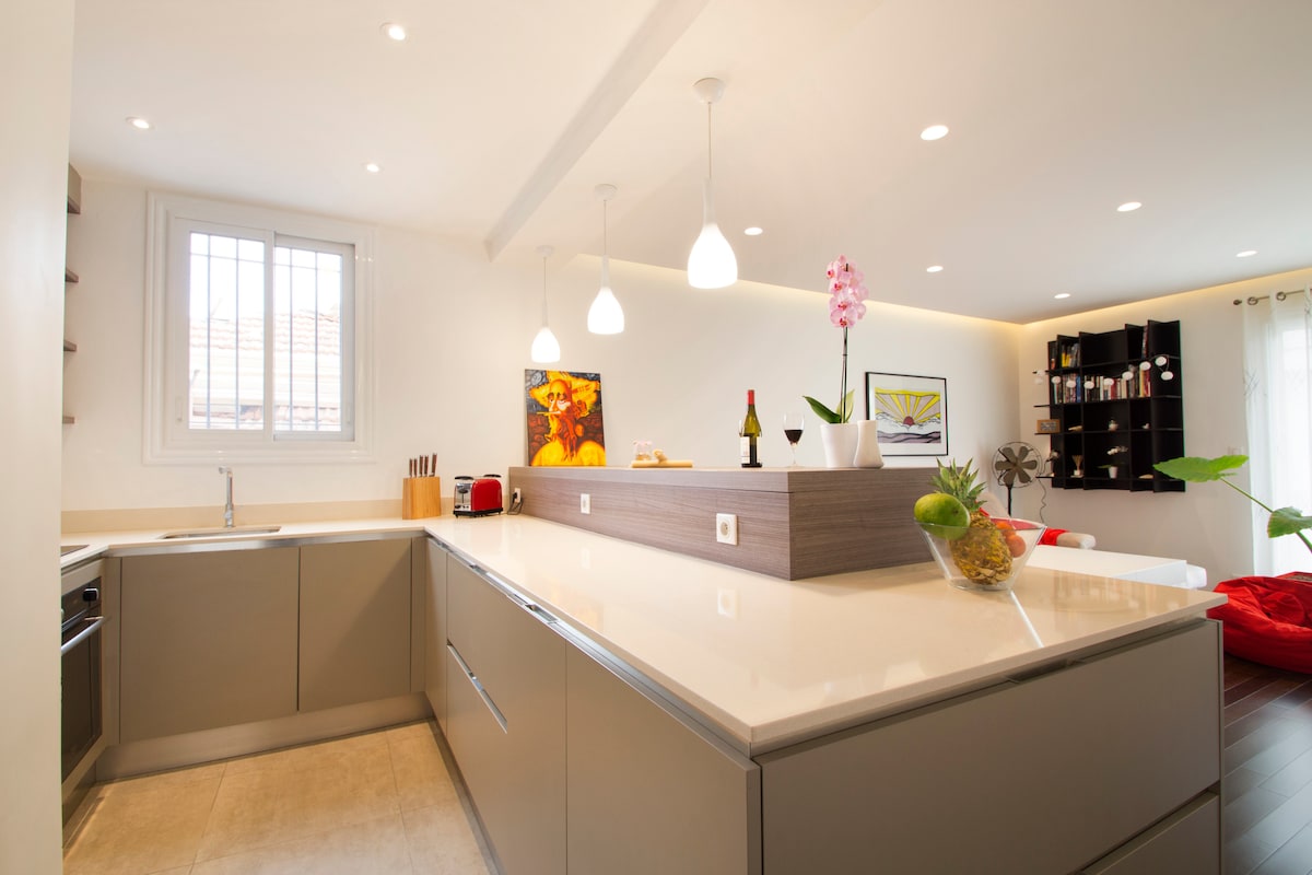 The kitchen features a modern design with sleek cabinetry and a spacious island. Under-cabinet lighting enhances the bright atmosphere, while pendant lights provide additional illumination. A large window allows natural light to fill the space, highlighting the various appliances and decorative plants.