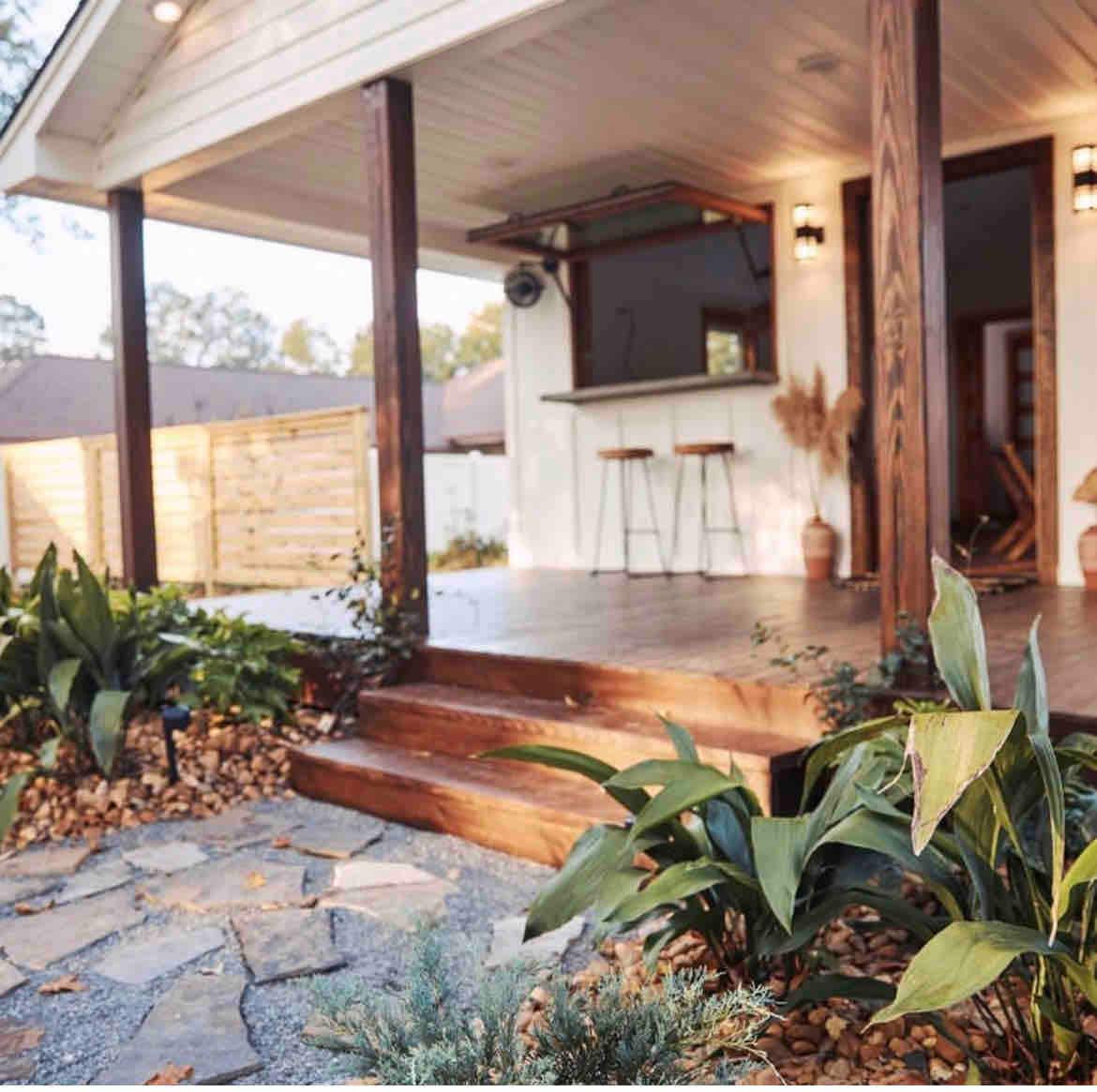 The inviting entrance features a wooden deck with steps leading up to a covered porch. Lush greenery and decorative pebbles accentuate the pathway, while natural wood pillars support the roof. A small bar area is visible, enhancing the outdoor living experience.