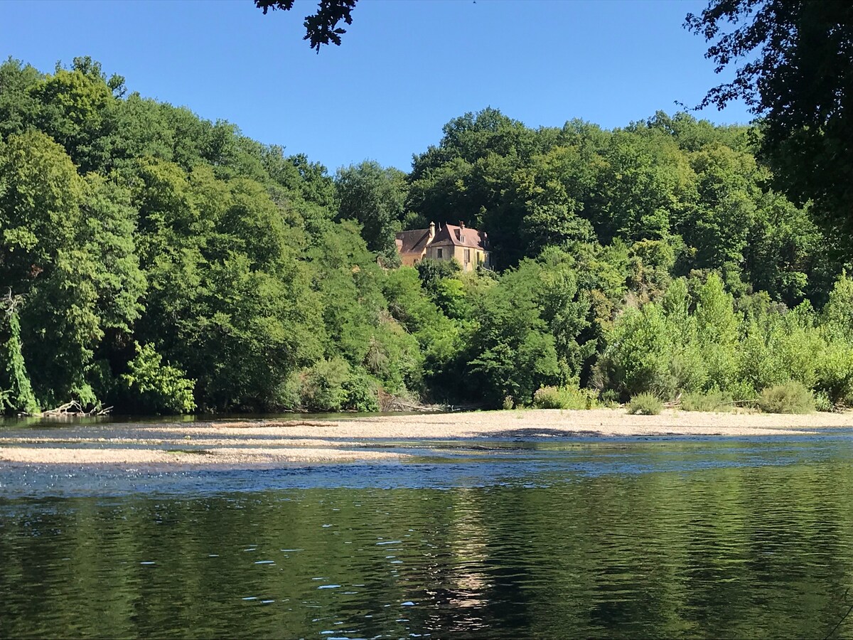 A traditional stone house is nestled among lush greenery on a hillside, overlooking a serene river. Clear blue skies reflect on the water, while the surrounding trees create a natural frame, enhancing the peaceful ambiance of the location.
