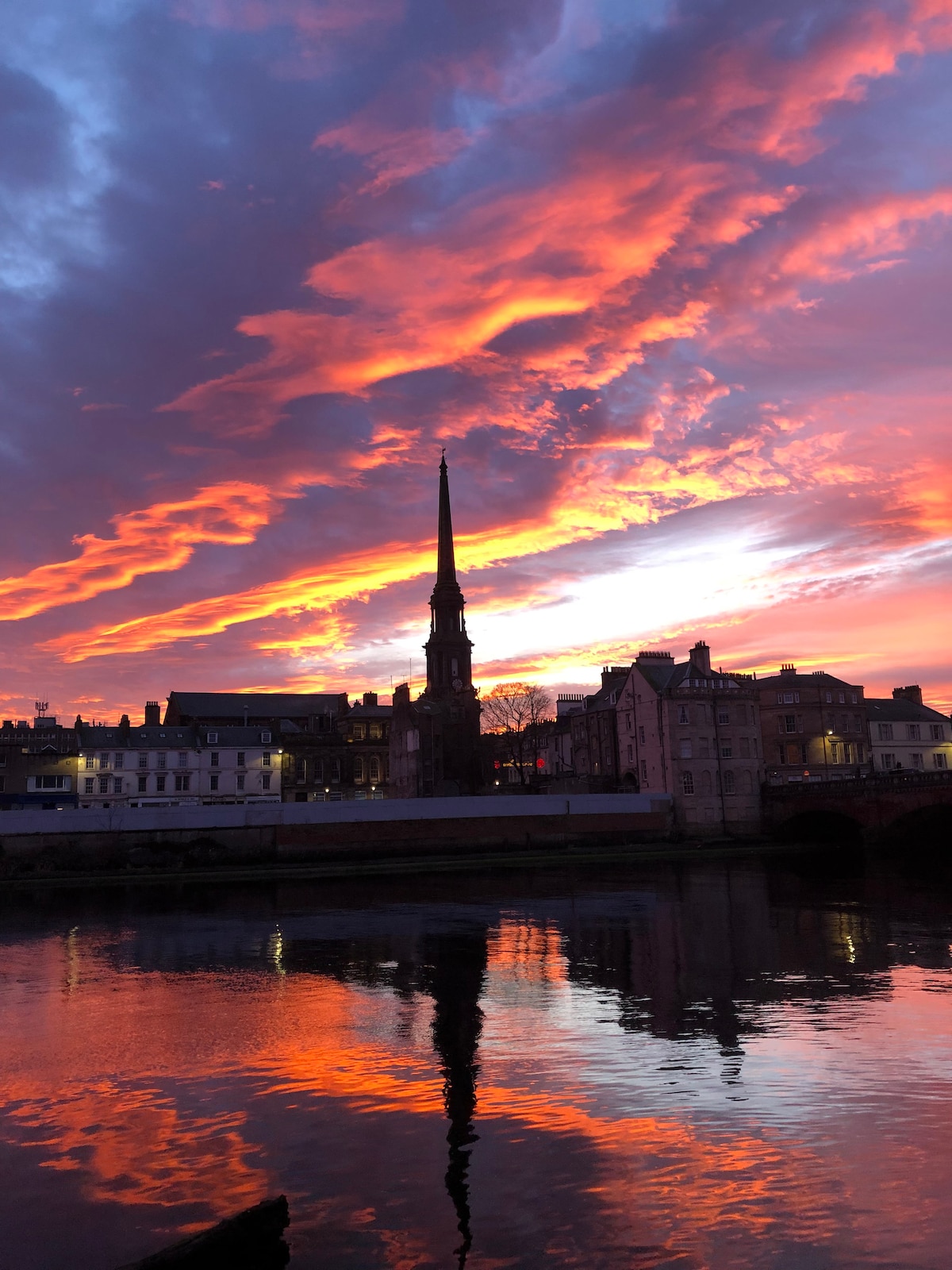 A vibrant sunset casts warm hues of orange and pink across the sky, reflecting on the calm river surface. A prominent church steeple rises against the backdrop of historic buildings, creating a scenic silhouette against the colorful sky.
