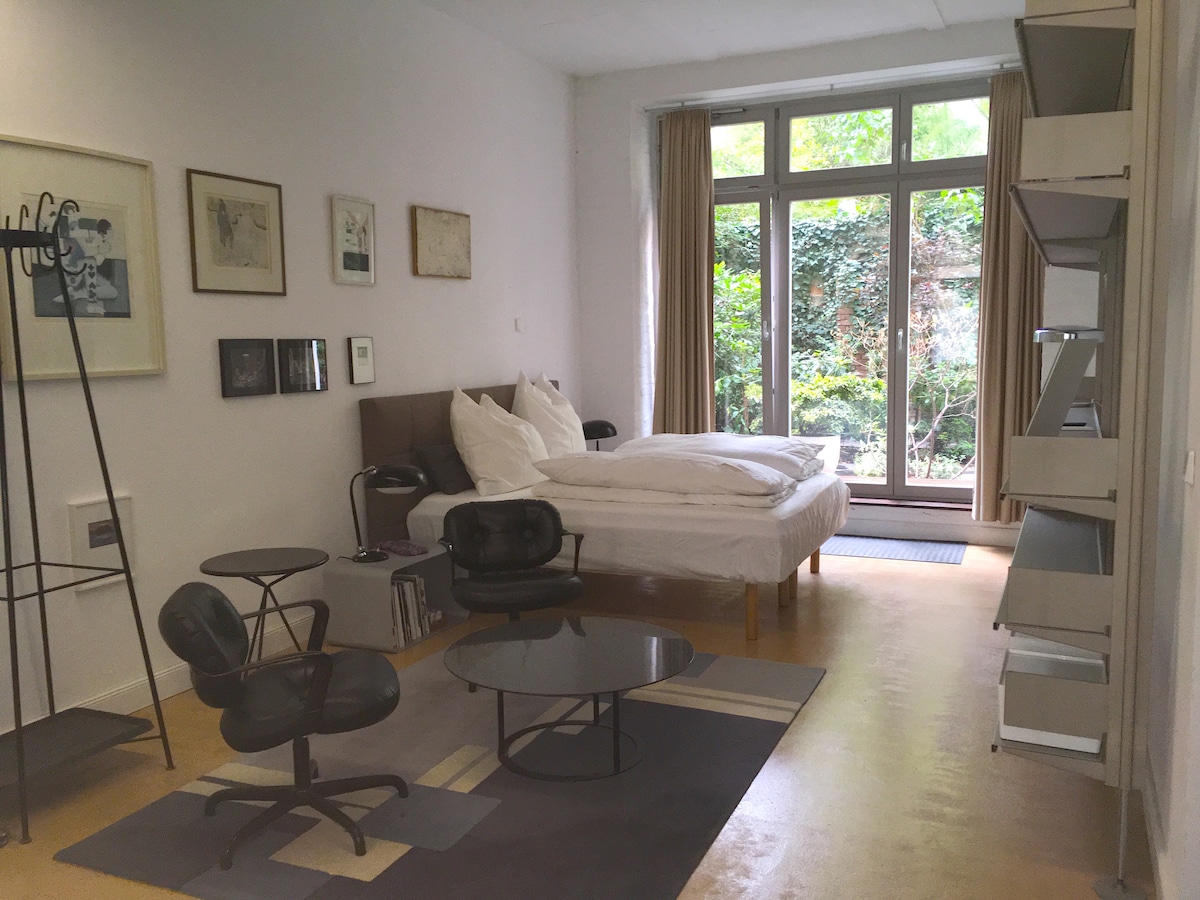 A spacious loft studio features a large bed with crisp white linens. Large windows provide natural light and a view of greenery outside. Mid-century furniture, including stylish chairs and a round coffee table, adds to the modern aesthetic. Art pieces adorn the walls.