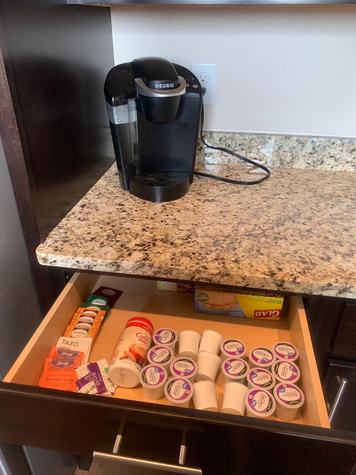 A coffee maker is positioned on a granite countertop beside a neatly organized drawer. The drawer contains various coffee pods and supplies, including creamers and sugar, suggesting convenient access to hot beverages.