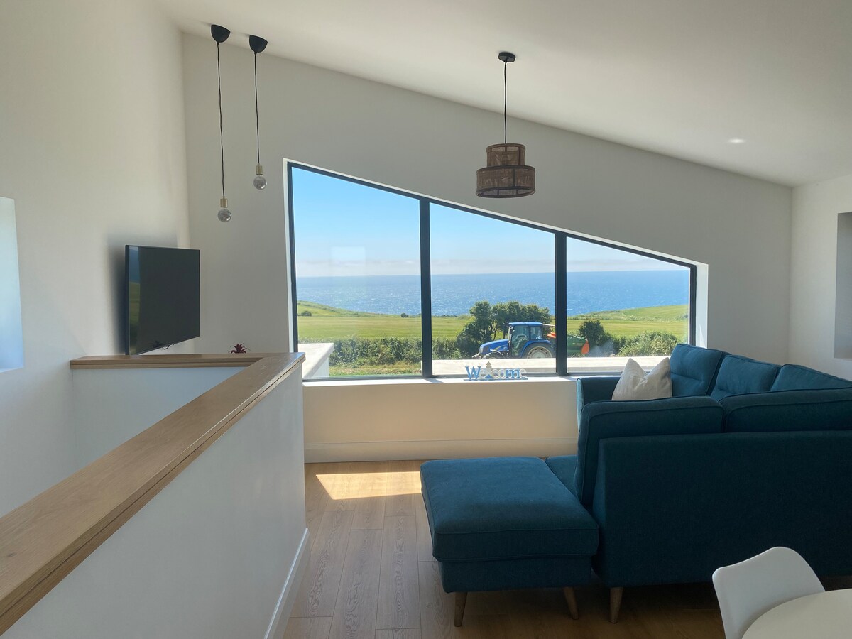 A bright living area features a segmented floor-to-ceiling window providing sweeping views of the sea and surrounding landscape. A cozy blue couch is positioned towards the window, enhancing the view. A mounted television is visible on the opposite wall, with minimalistic design elements throughout.