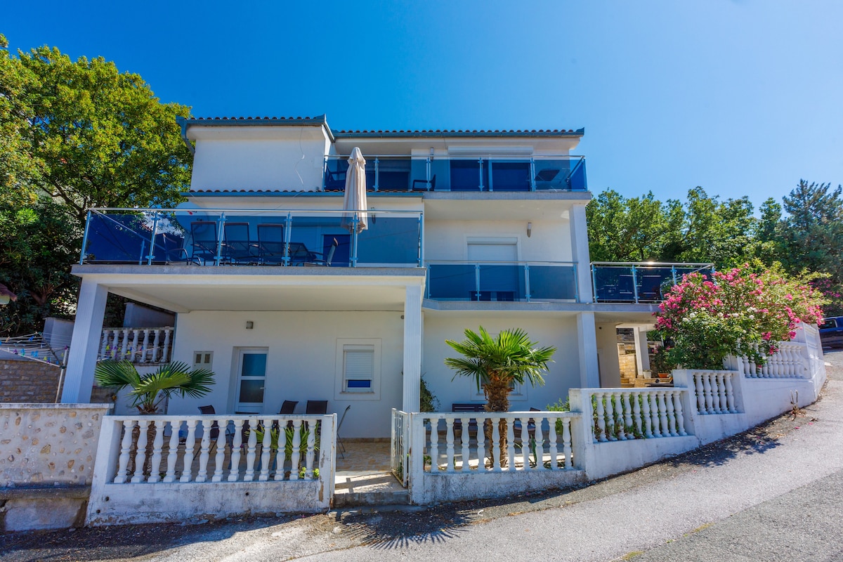 A modern multi-level villa is showcased, surrounded by greenery. The façade features large balconies with glass railings, offering outdoor seating options. Tropical plants and flowering shrubs are visible in the garden area, creating an inviting entrance to the home.