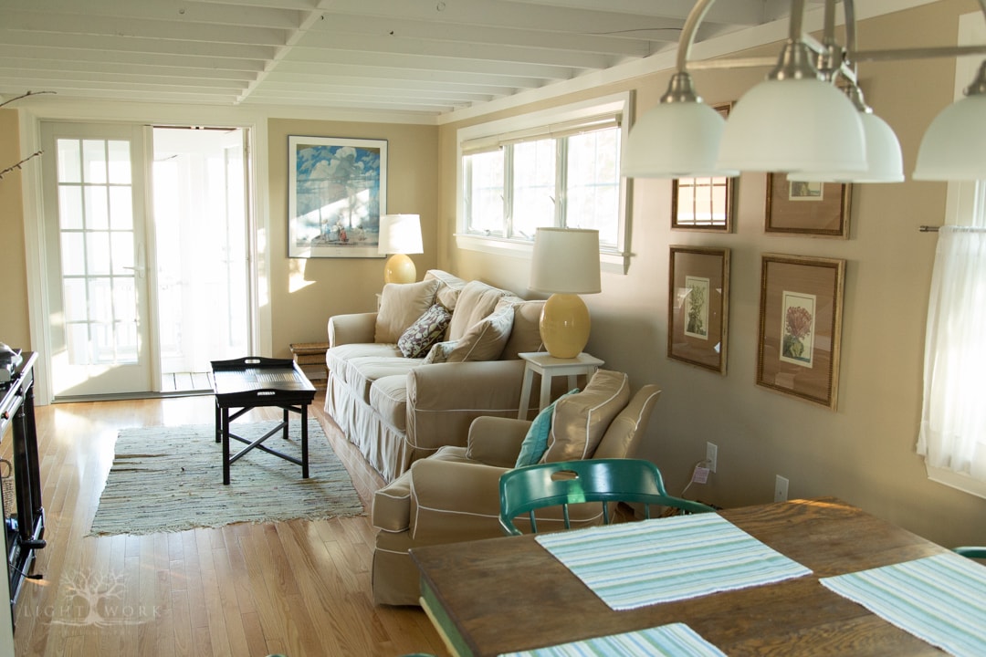 A cozy living area offers a light-filled space with a comfortable sofa and armchair. A wooden coffee table is positioned in front of the seating. The adjacent dining area features a wooden table with two chairs, welcoming natural light from multiple windows.