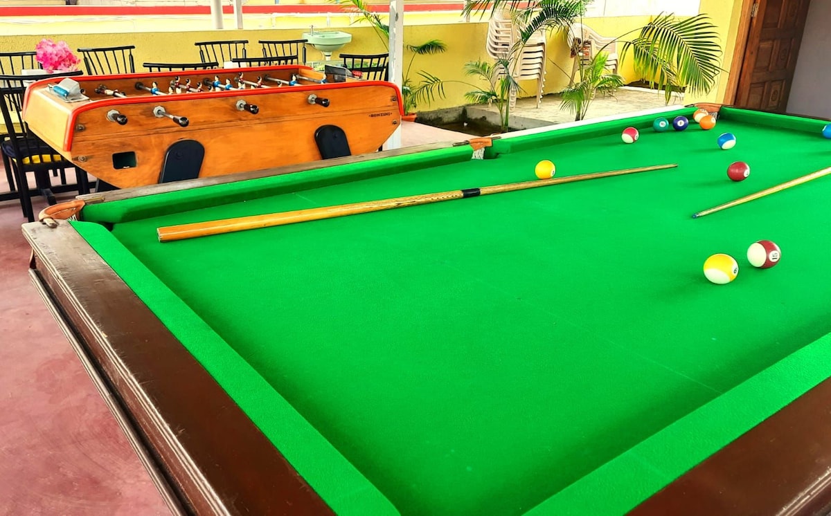 A vibrant gaming area features a green pool table and a foosball table, both positioned on a smooth surface. Various colorful balls are scattered across the pool table, and chairs are visible in the background, enhancing the entertainment space.
