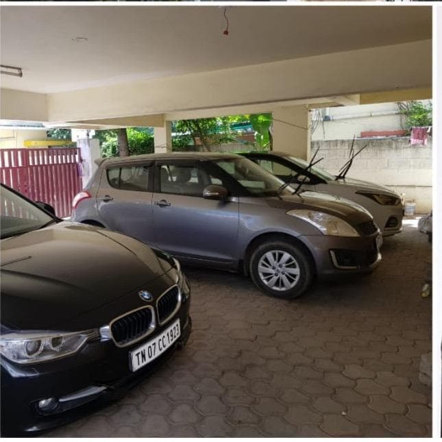 A covered parking space accommodates three vehicles, featuring a black BMW and two other cars in muted tones. The space is outlined by a low fence, with greenery visible in the background, providing a sense of privacy and seclusion.
