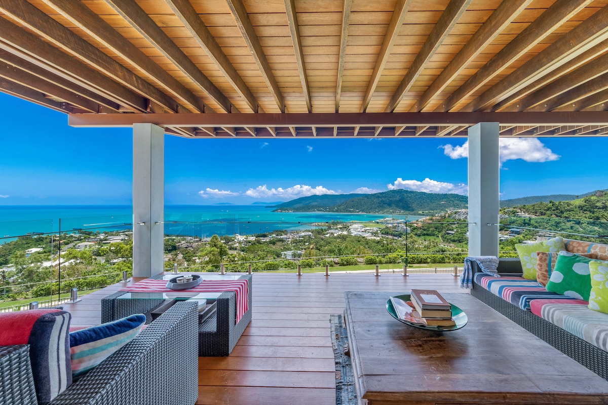 An outdoor seating area is positioned under a wooden overhang, featuring comfortable wicker furniture and colorful cushions. A large coffee table holds books, while stunning panoramic views of the Whitsundays Islands and Airlie Beach are visible in the background.