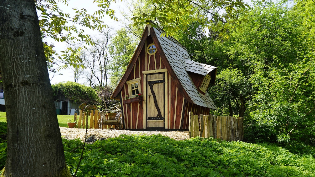 A charming wooden cabin features a sloped roof and a rustic door, set amidst lush greenery. Its windows, adorned with shutters, add character. A small outdoor seating area is visible, inviting guests to enjoy the surrounding natural beauty.