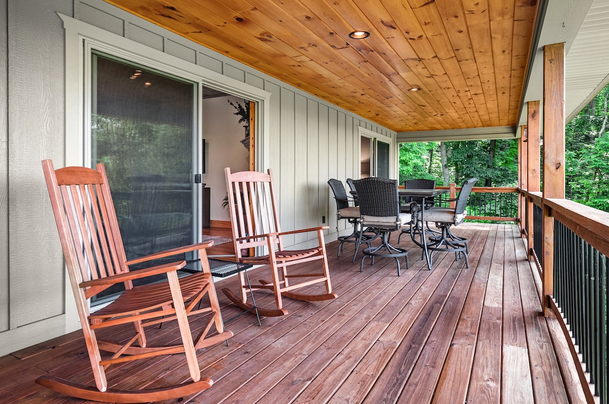 A covered deck features wooden rocking chairs paired with a dining set. The warm wood ceiling complements the natural tones of the wooden deck. Lush greenery is visible in the background, enhancing the sense of outdoor space.