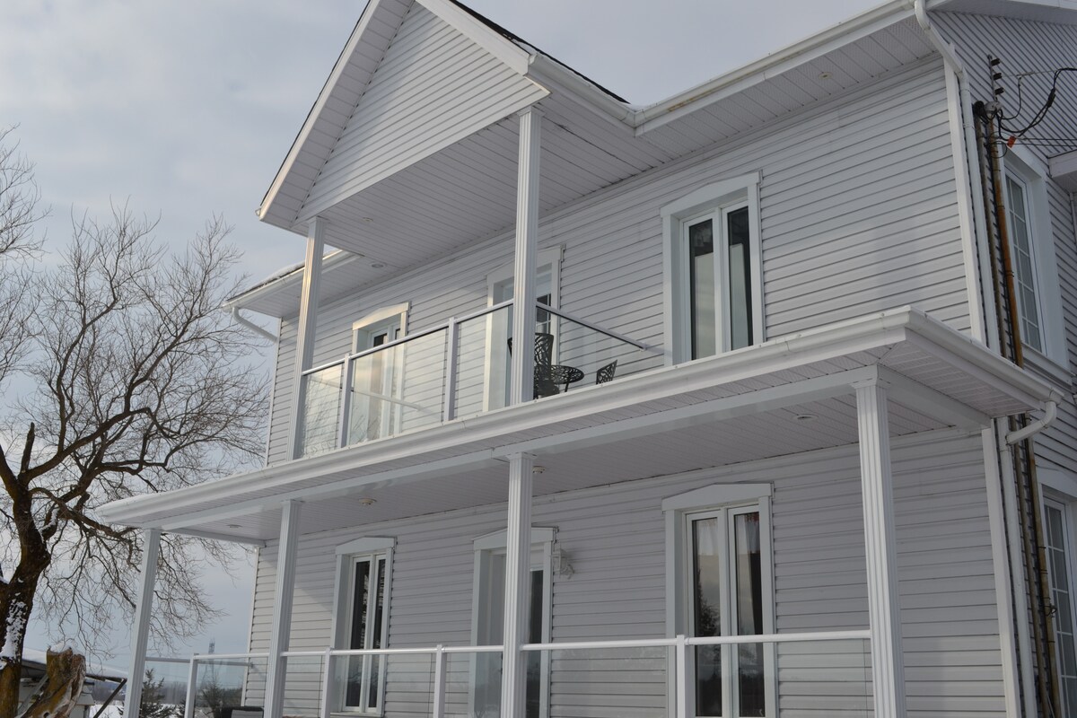 The exterior of the house is shown, featuring a light-colored facade with multiple windows. A second-floor balcony is visible, supported by white columns, and furnished with a small table and chairs. A tree stands in the foreground, adding to the rural ambiance.