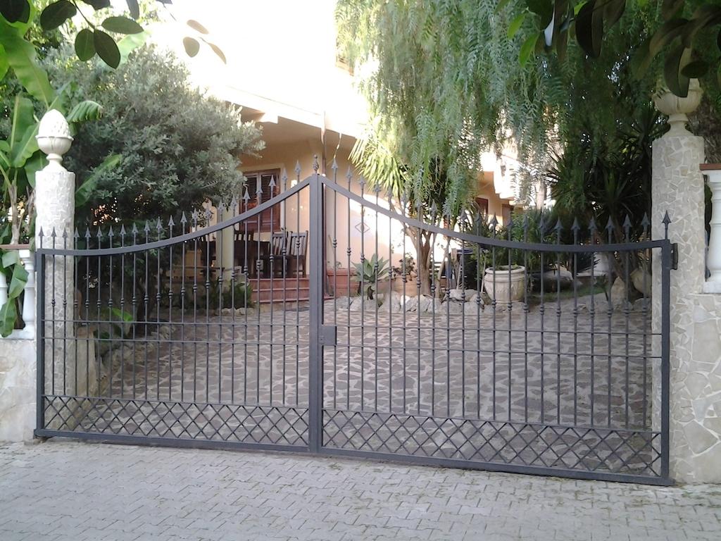 An ornamental wrought iron gate is shown, leading to a landscaped pathway. Lush greenery surrounds the entrance, and a set of steps is visible in the background, leading to a home with a covered porch and seating.