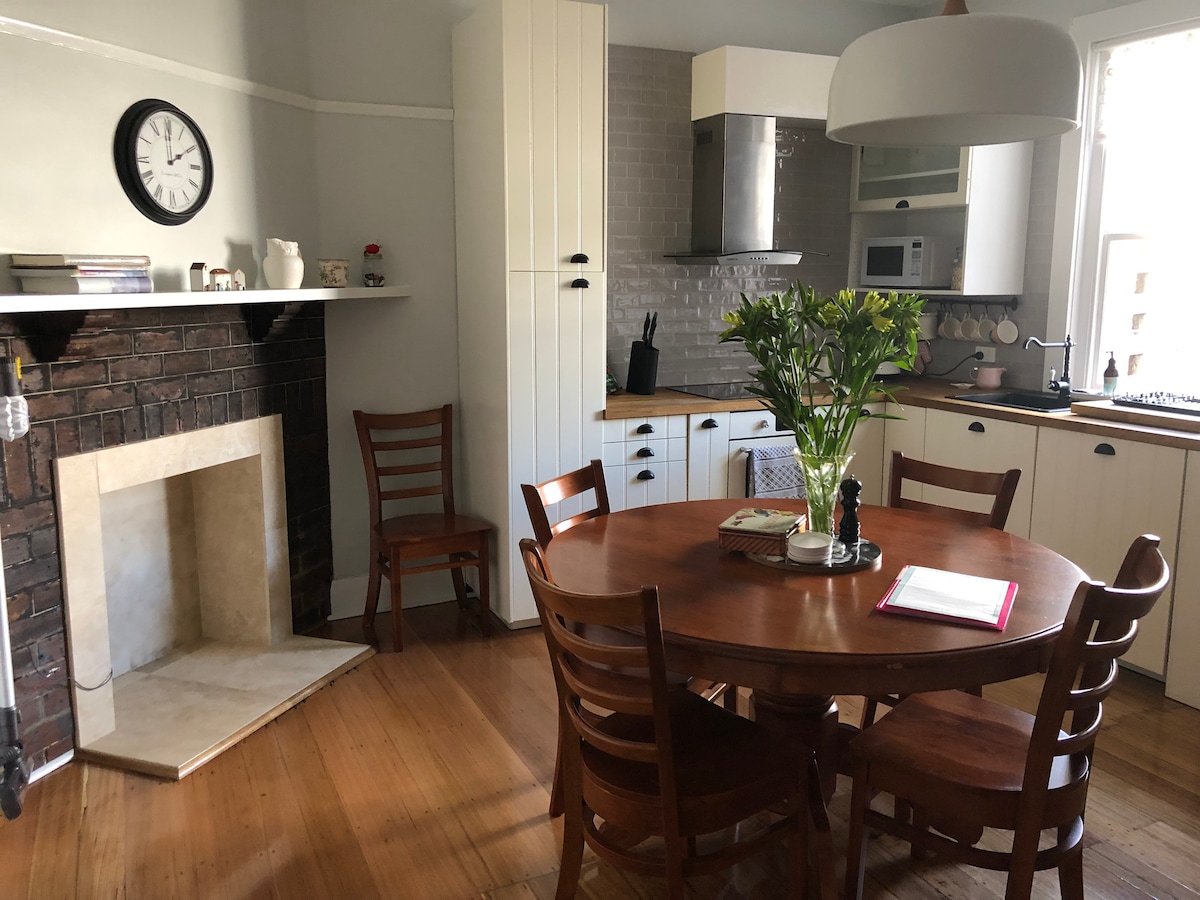 A well-organized kitchen and dining area features a round wooden table surrounded by four chairs. The kitchen includes white cabinetry and modern appliances, with a fireplace adding a vintage touch. Fresh flowers are arranged on the table, enhancing the inviting atmosphere.