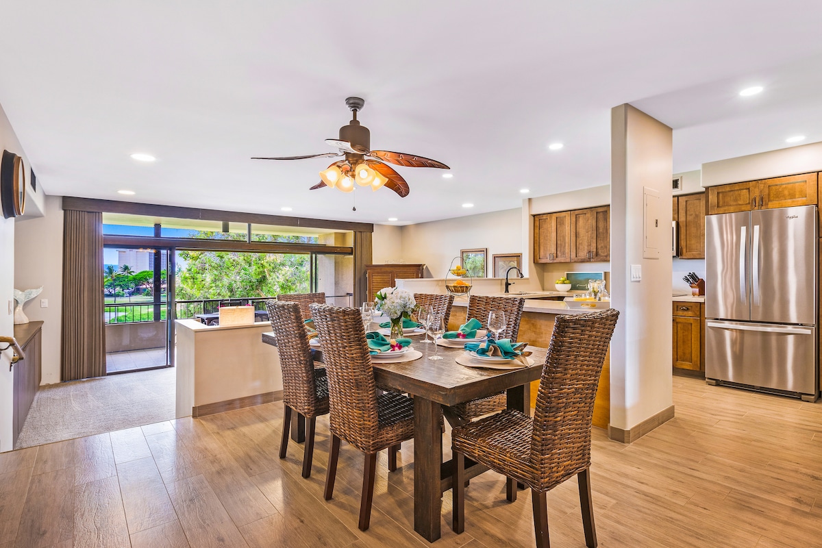 A spacious dining area includes a dark wooden table set for six, surrounded by woven chairs. The kitchen is visible in the background, featuring modern stainless steel appliances and warm wooden cabinetry. Large windows invite natural light, offering views of the lush outdoor landscape.