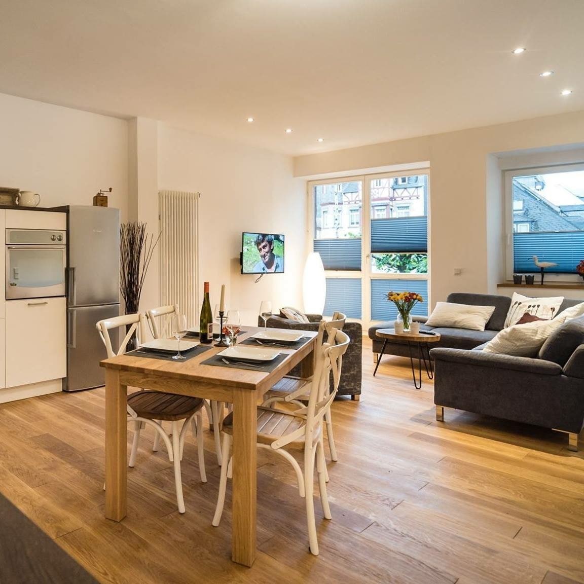 A modern dining area features a wooden table set for four, accompanied by light-colored chairs. A gray sofa sits nearby, with a television mounted on the wall. Natural light enters through large windows, illuminating the open layout.
