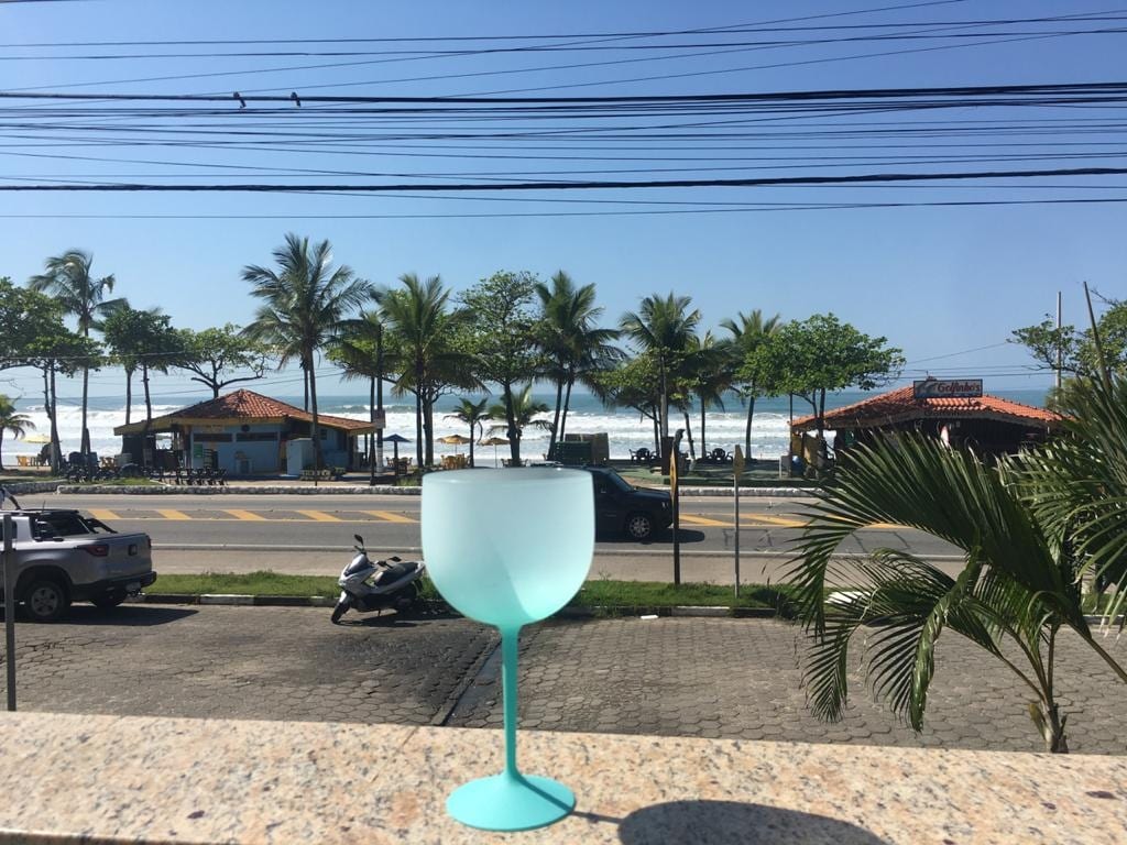 A scenic view from a balcony is captured, displaying a vibrant coastline. Palms line the shore, with beachside establishments visible in the distance. The ocean waves can be seen rolling onto the sandy beach, while a teal glass stands prominently in the foreground.