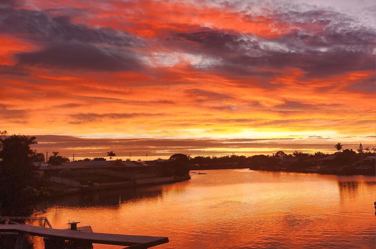 A vibrant sunset illuminates the sky with shades of orange, red, and purple, reflecting onto the calm water below. Silhouettes of trees and homes line the horizon, creating a serene backdrop to the natural display of colors.