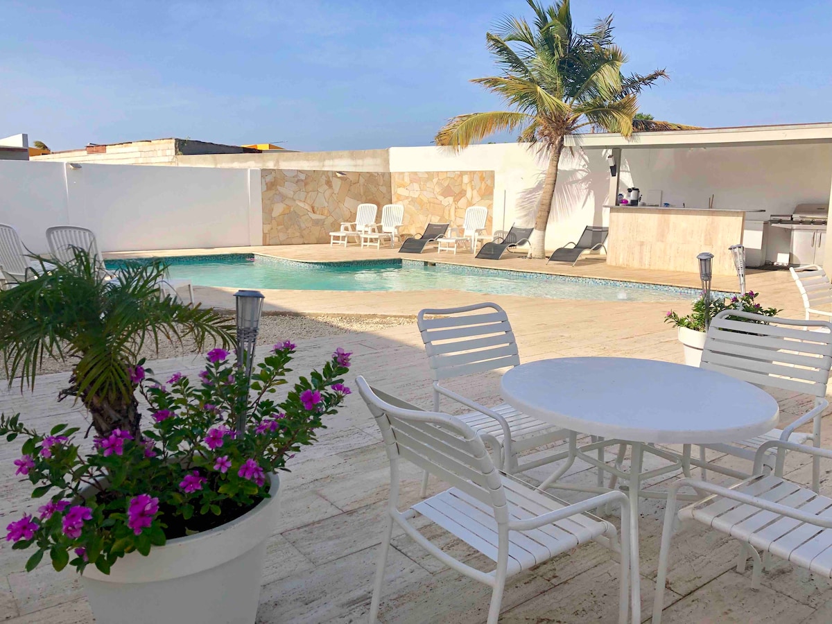 A serene outdoor area features a clear swimming pool bordered by sun loungers and a shaded seating area. Potted plants with vibrant flowers add color, while a modern outdoor kitchen area is visible in the background, set against a bright blue sky.