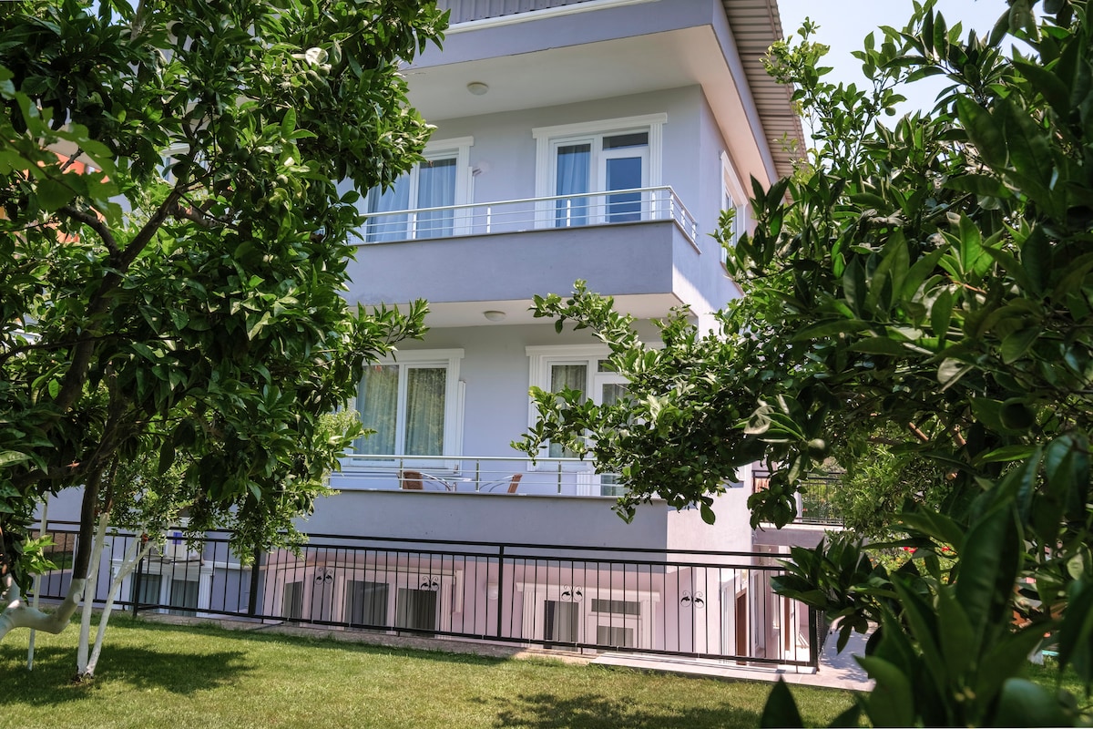 The exterior of a two-story building is framed by lush greenery, including orange trees. Multiple air conditioning units can be seen, and spacious balconies are visible with chairs placed for relaxation. The manicured lawn adds to the inviting atmosphere of the property.