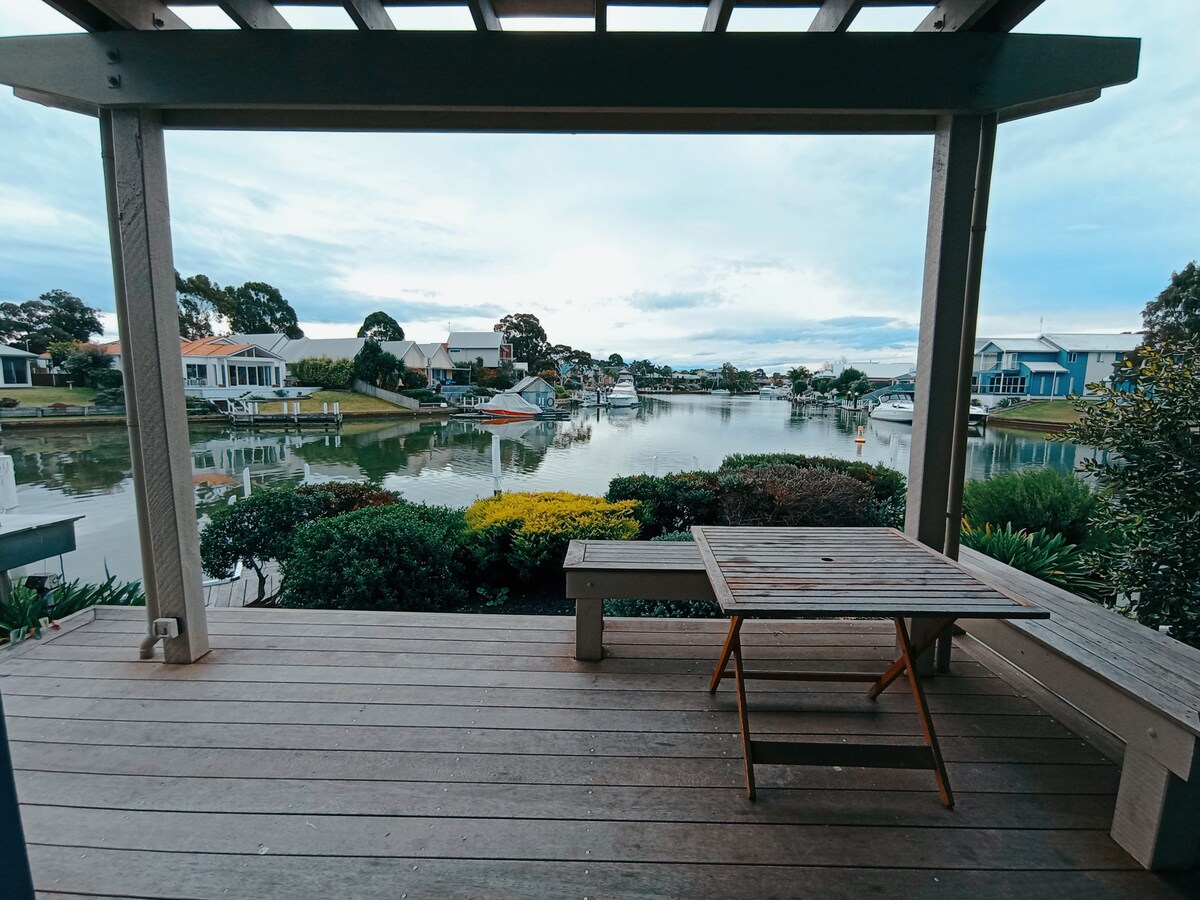 A spacious deck features a wooden table, overlooking a serene waterfront view. The area is surrounded by greenery, with various boats moored nearby. The calm water reflects the sky, creating a tranquil atmosphere.