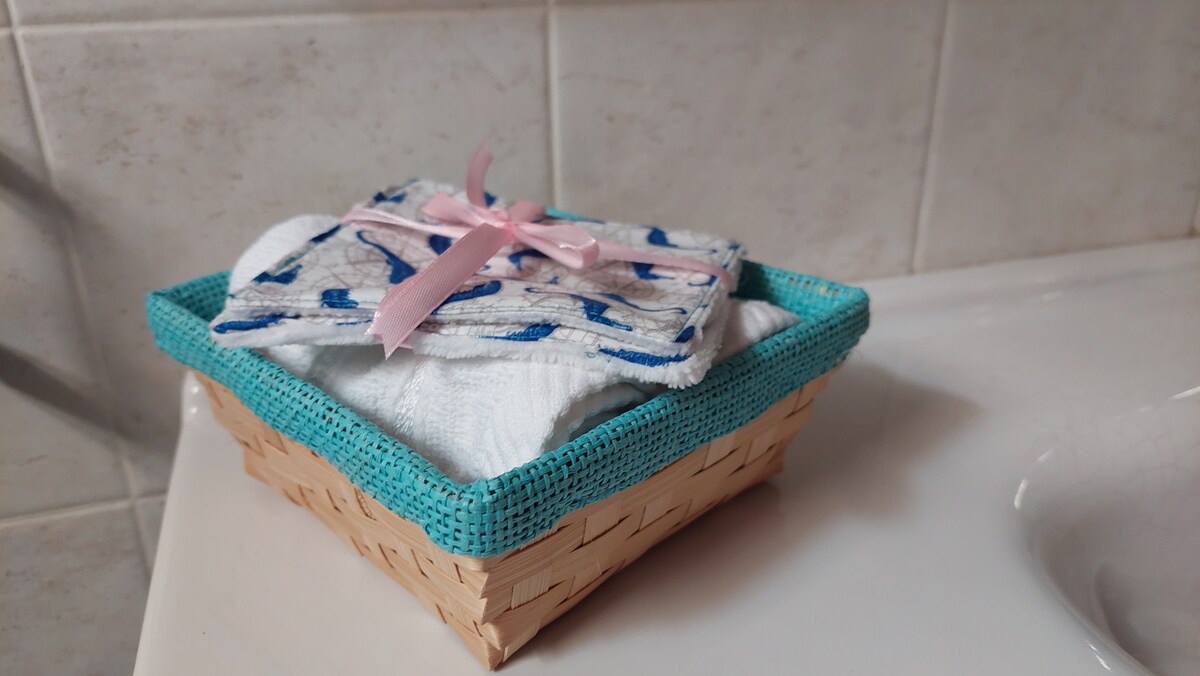 A woven basket with a turquoise border holds soft, neatly stacked towels and two decorative cloths tied with a pink ribbon. The basket is positioned on a countertop, against a light-colored tiled wall.