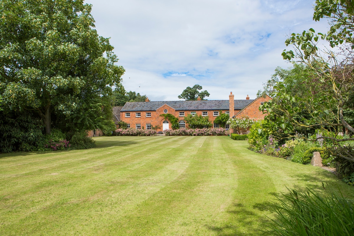 A spacious lawn leads to a charming brick building surrounded by lush greenery. Mature trees and colorful flower beds frame the property, creating a serene outdoor setting. The sky is partly cloudy, contributing to a peaceful atmosphere at the farm.