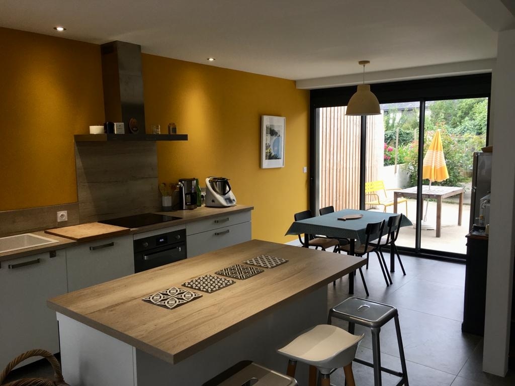 An open kitchen area features modern cabinetry and a wooden countertop. A dining table is positioned nearby, with metal stools arranged around it. Large glass doors lead to an outdoor courtyard, which includes a colorful umbrella and seating options.