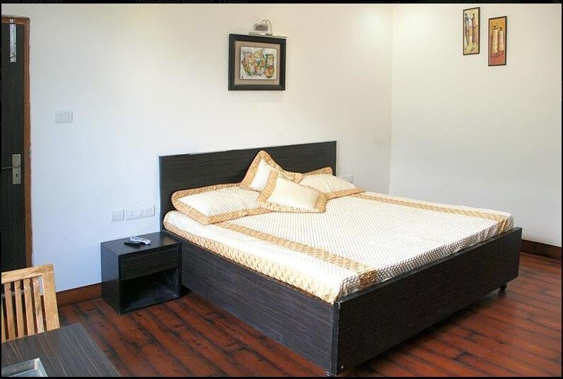 A spacious guest room features a large bed adorned with golden accents and complementary pillows. Light wooden flooring enhances the modern design. A bedside table is placed next to the bed, while a wooden dining table is visible in the corner, offering a comfortable space for relaxation.