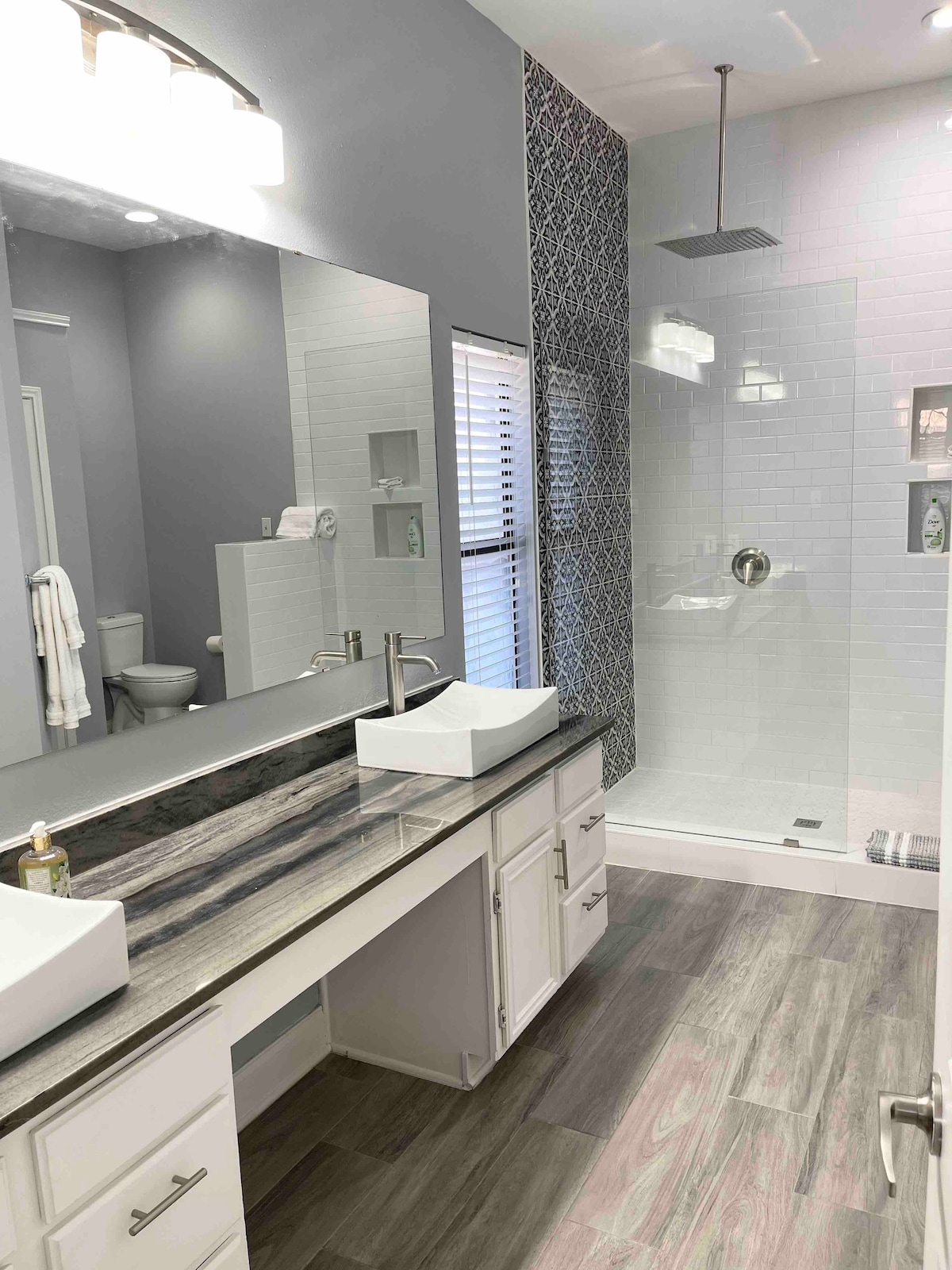 A modern bathroom features dual sinks with stylish vessel sinks set on a dark countertop. The walk-in shower is enclosed in clear glass, showcasing a rainfall showerhead. Natural light is allowed in through a window with blinds, and neutral tones create a clean and spacious feel.