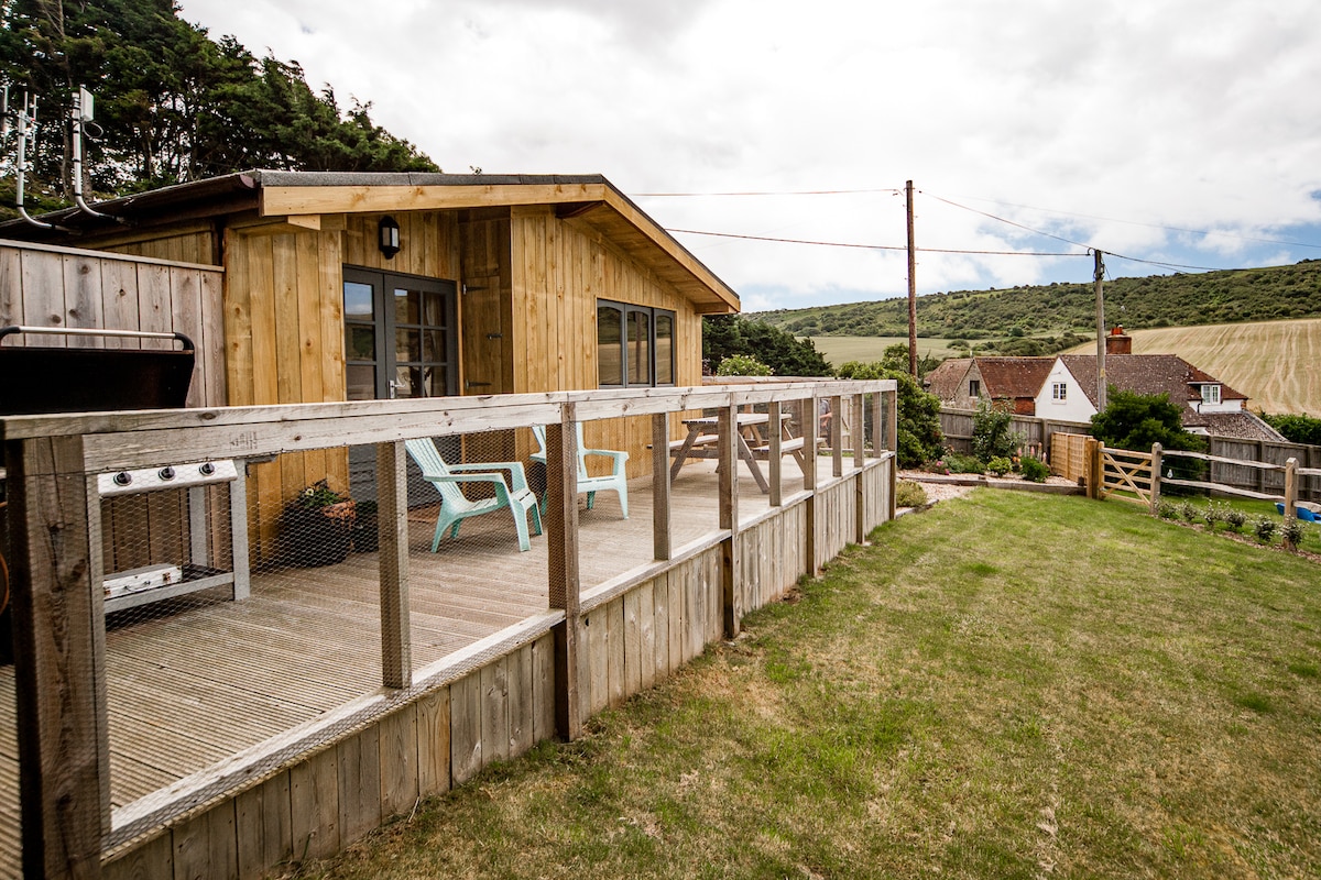 A wooden cabin is set on a spacious deck with seating, providing a relaxed outdoor space. The grassy area extends towards the surrounding countryside, showcasing a blend of natural beauty and rural charm. The cabin's exterior features natural wood tones and large windows.