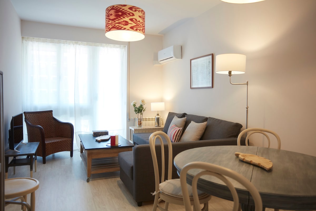 A comfortable living area is visible, featuring a gray sofa adorned with decorative pillows. A wooden coffee table sits in front, accompanied by a woven chair. Light streams through large windows, with soft curtains filtering the view. A dining table and decorative lamps complete the inviting space.