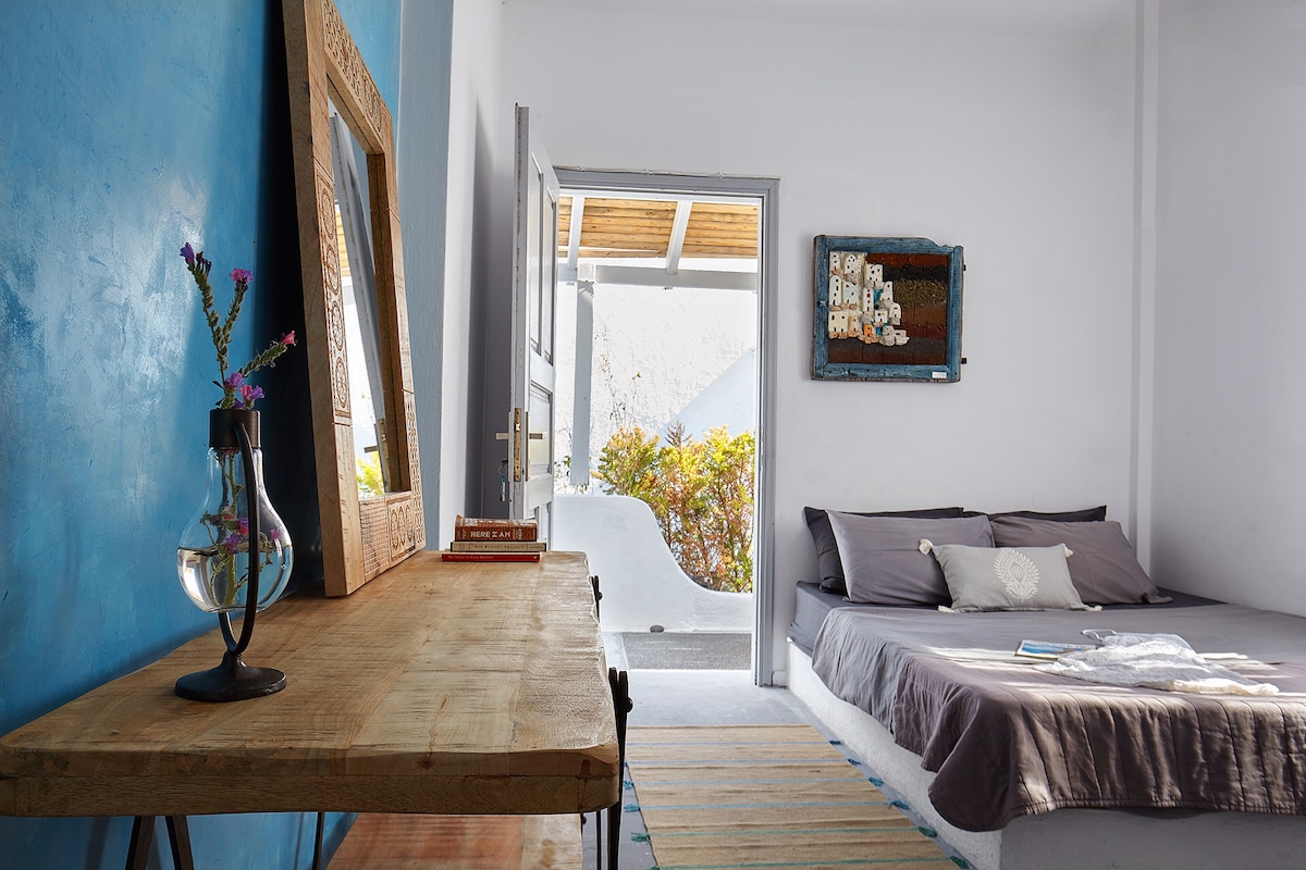A bright, airy room showcases a spacious bed with subtle gray bedding against a calming blue wall. A wooden table with a lamp is positioned nearby, complemented by a mirror and artwork on the wall. The open door leads to a serene outdoor space.
