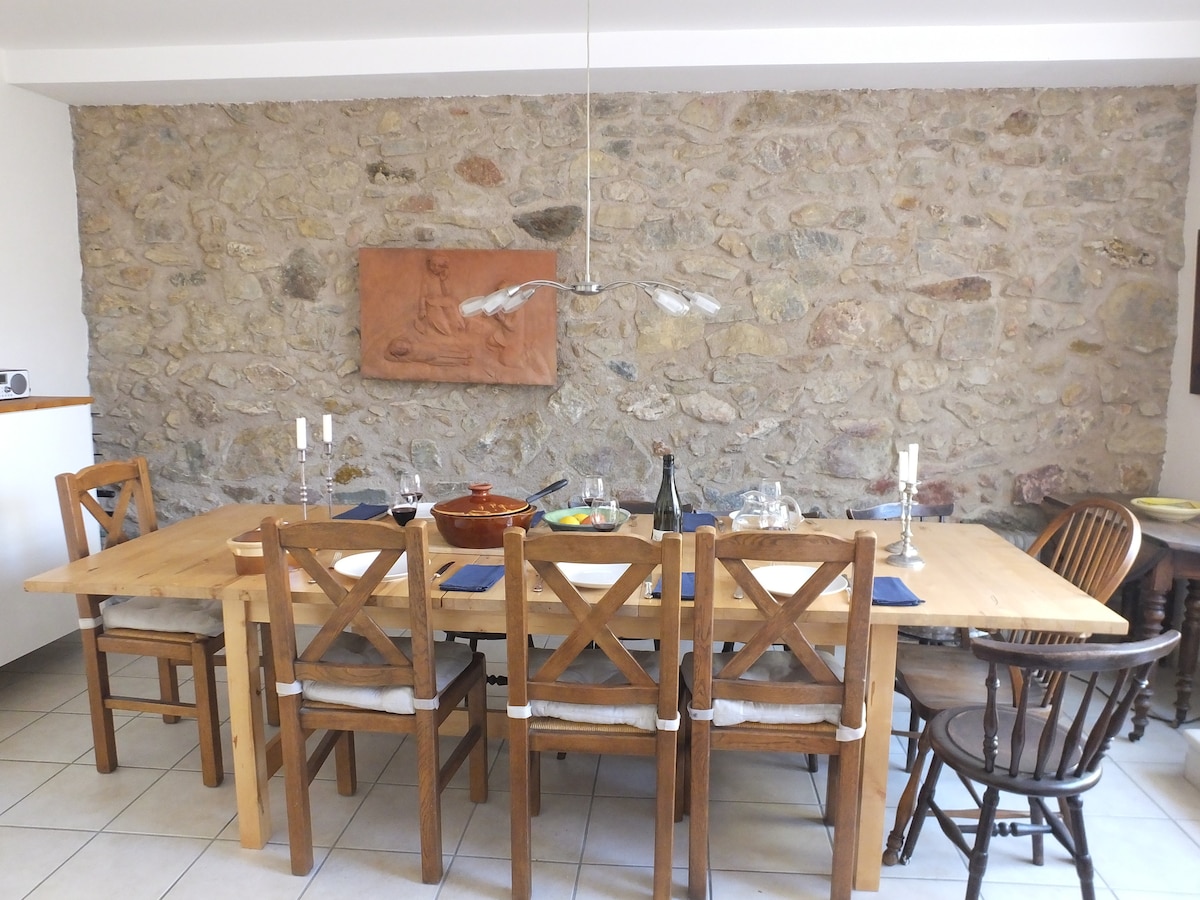 A spacious dining area is presented with a wooden table surrounded by six chairs, featuring a mix of styles. The stone wall adds texture, while a decorative art piece hangs above the table. Simple table settings are arranged with plates and utensils.