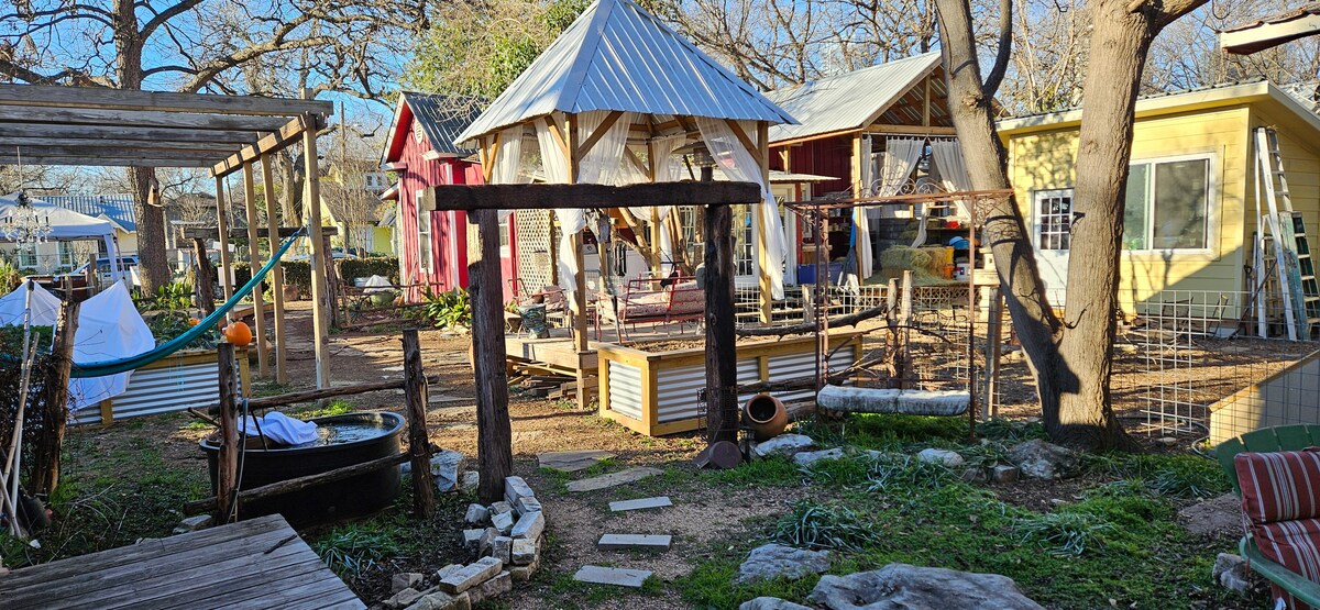 A rustic outdoor space is captured, featuring a mix of colorful structures including a gazebo and small cottages. A stone pathway leads through a garden area, surrounded by trees and open space. Hammocks and outdoor furniture are arranged for relaxation.