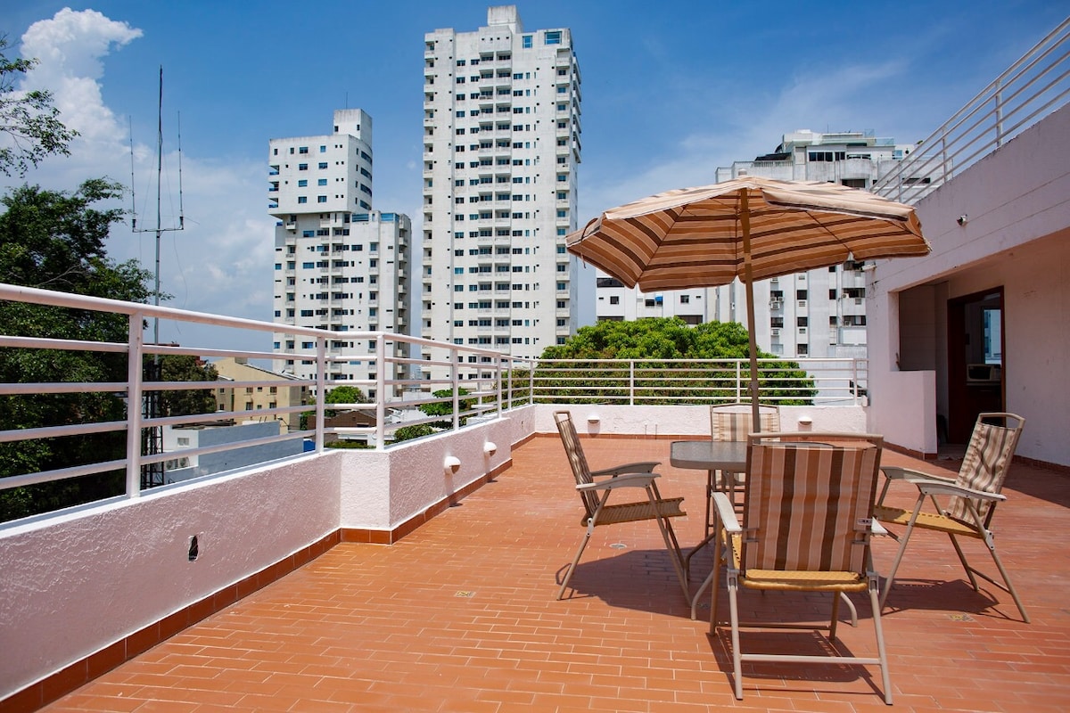 A spacious rooftop terrace is showcased, featuring a large table surrounded by four striped chairs. An umbrella offers shade, while views of surrounding tall buildings and blue skies create a pleasant outdoor setting.