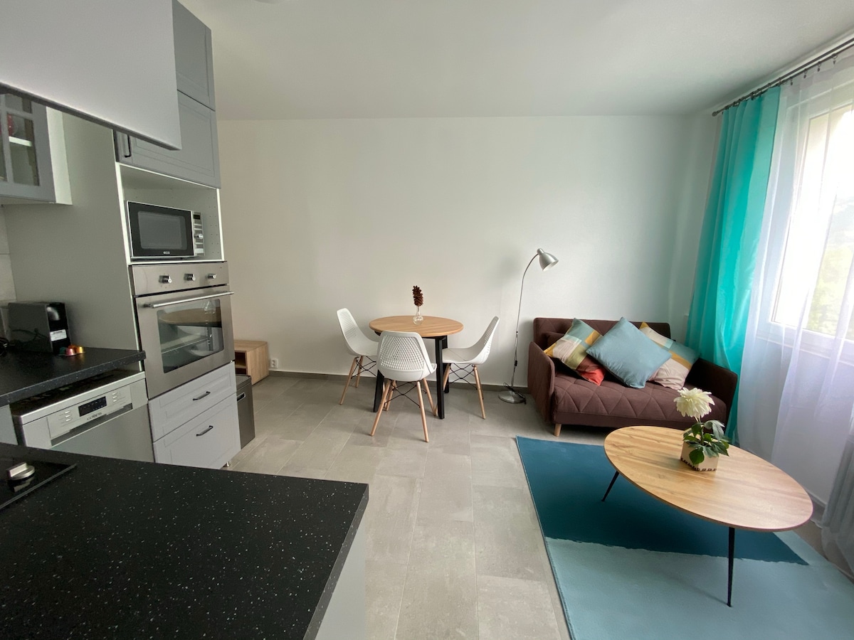 A modern open-plan living space features a comfortable brown sofa adorned with colorful cushions, a round dining table with white chairs, and a stylish coffee table. Natural light filters through the window, enhancing the bright and airy atmosphere.