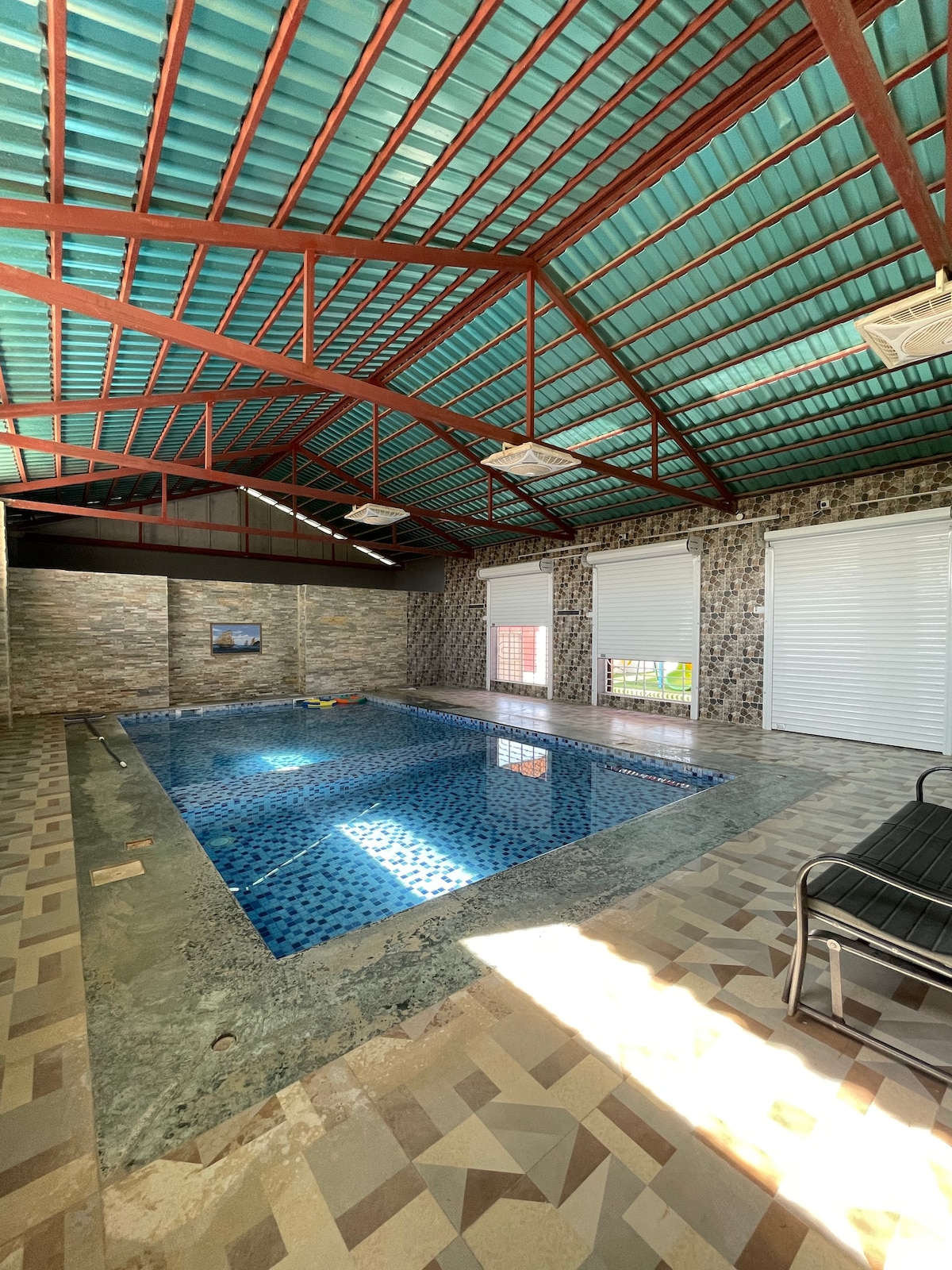 An indoor swimming pool is featured with azure tiles reflecting the light from overhead windows. The spacious area is surrounded by stone walls and a polished floor, while a few lounge chairs are positioned nearby for relaxation.