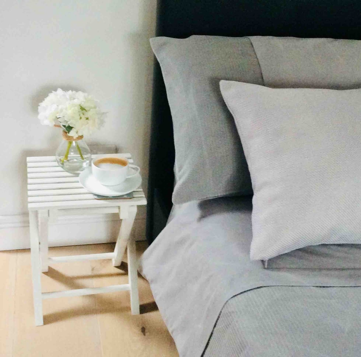 A cozy corner of the room features a spacious bed with soft grey linens. A small white table holds a cup of tea and a vase of delicate flowers, providing a serene and welcoming touch.