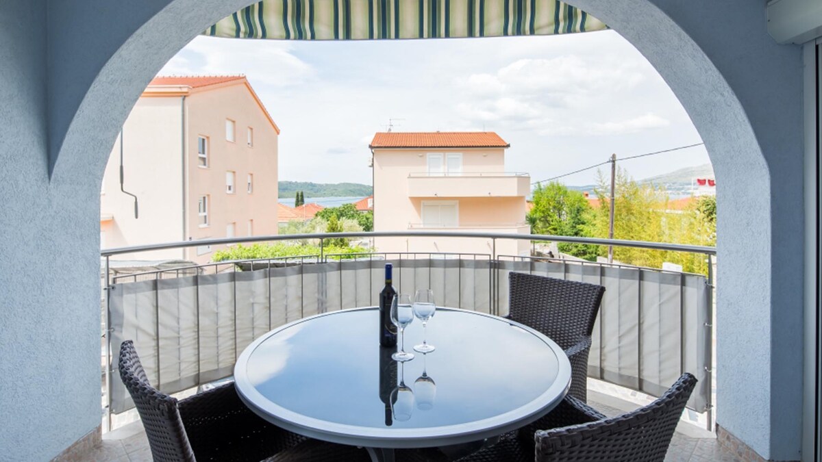 A balcony is highlighted by a round table surrounded by four wicker chairs. A bottle of wine and two glasses are elegantly placed on the table. The view includes nearby buildings and glimpses of greenery, framed by a gentle archway and an awning above.