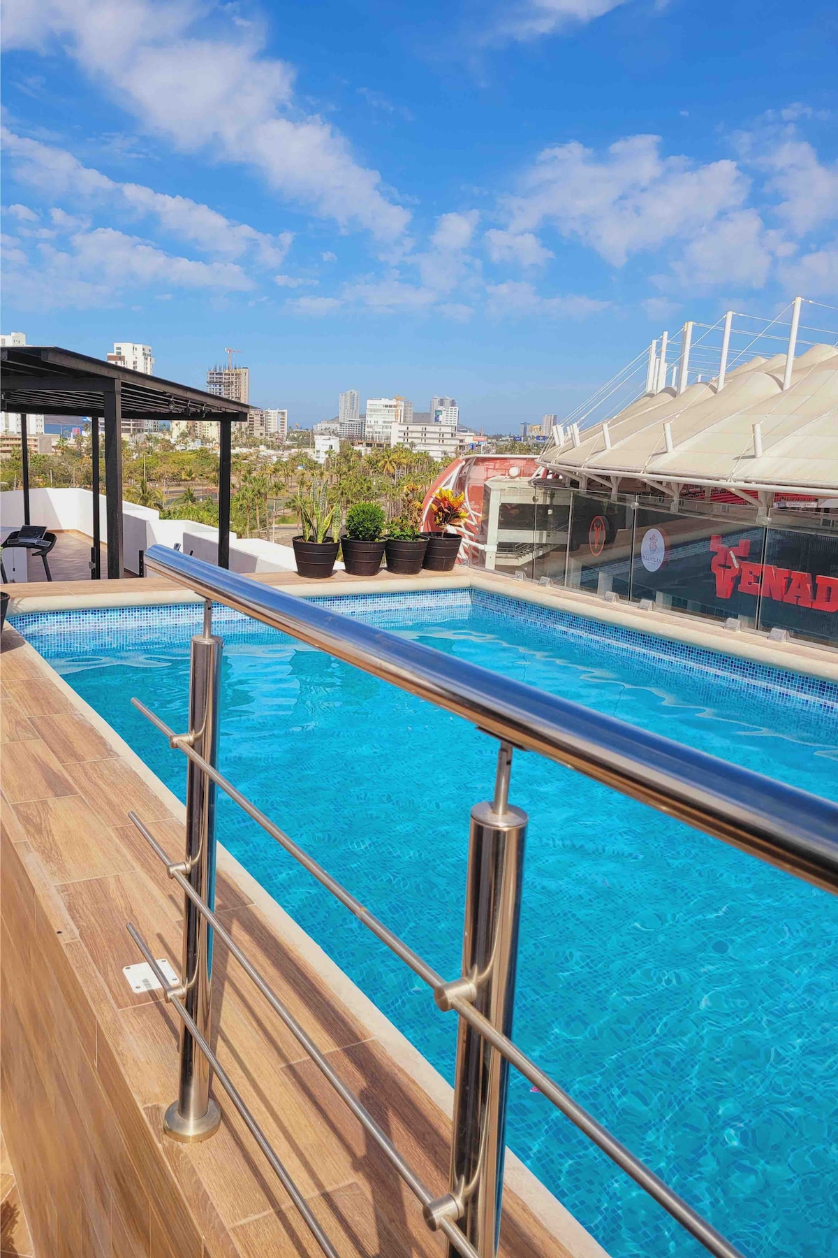 A rooftop pool is visible, featuring clear blue water under a bright sky. Surrounding the pool are landscaped planters with greenery, and the urban skyline can be seen in the distance. A shaded seating area is included, offering a place for relaxation.
