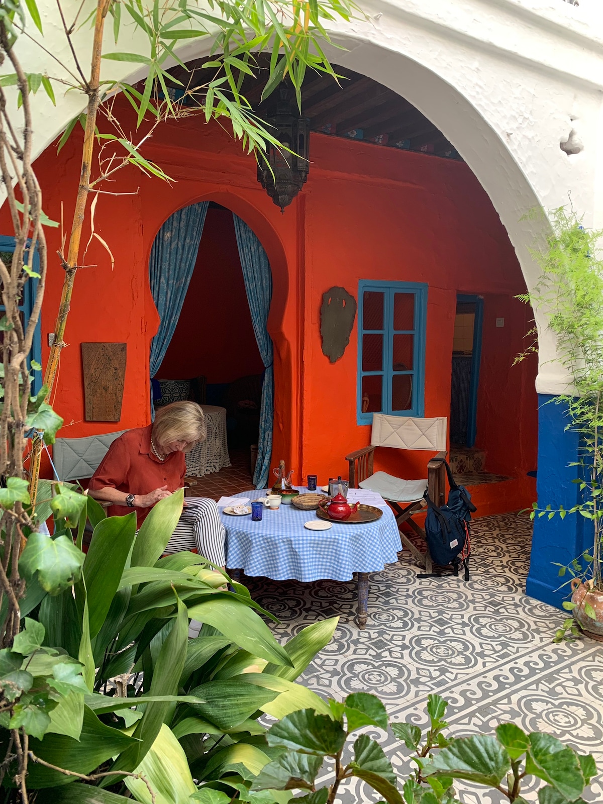 A vibrant courtyard is framed by lush greenery. An intricately patterned tile floor complements the brightly painted orange wall. A small blue table is set with dishes and drinks, and two chairs are positioned nearby. A person sits at the table, engaged in their activities.