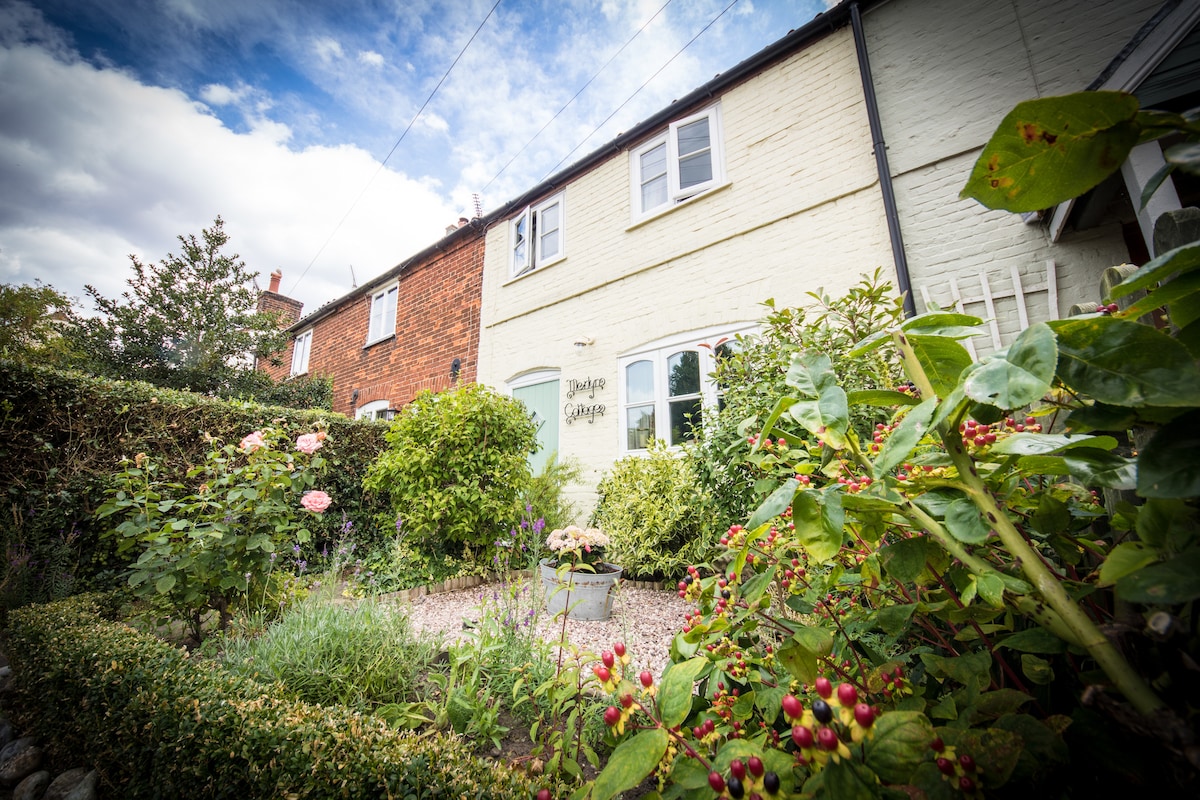 A charming cottage exterior is framed by a flourishing garden, featuring an array of colorful flowers and shrubs. The inviting entrance is marked by a sign, with light-colored walls contrasting against the greenery. A blue sky dotted with clouds provides a serene backdrop.