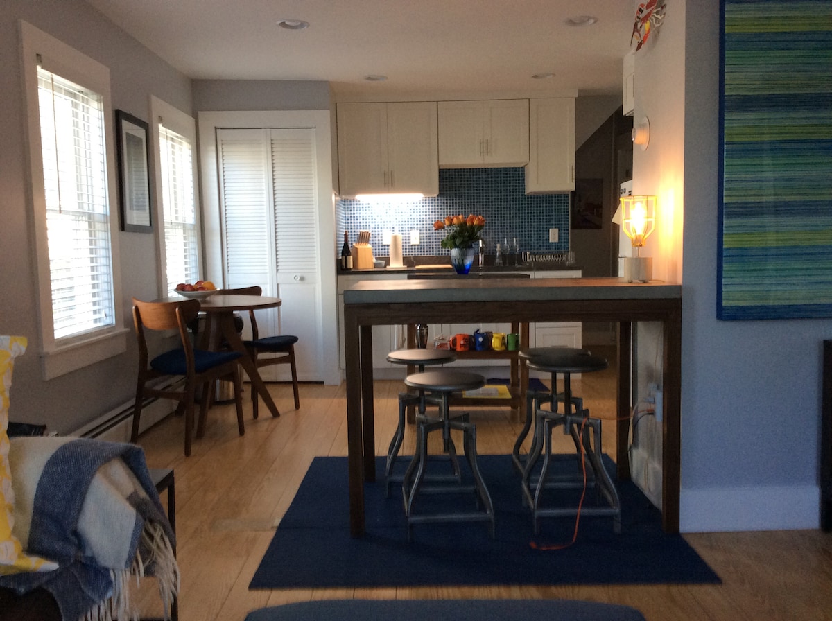 The image showcases a newly renovated kitchen and dining area with a rectangular wooden table and four chairs. A blue tiled backsplash is visible behind a modern countertop, accented by a vase of flowers. Natural light filters in through multiple windows.