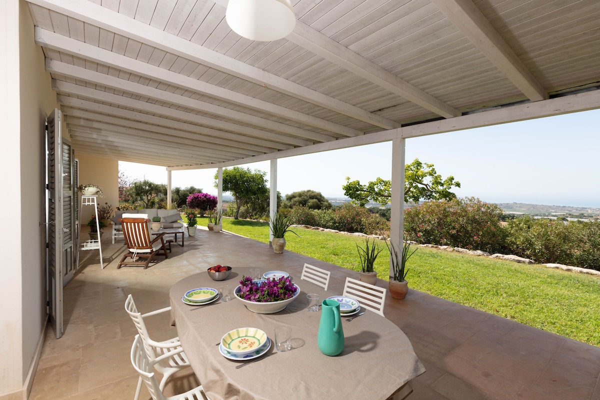 An outdoor terrace is furnished with a large dining table set for a meal, surrounded by white chairs. Potted plants and vibrant flowers accentuate the space, while comfortable seating areas invite relaxation. The open layout frames views of the lush countryside and distant sea.