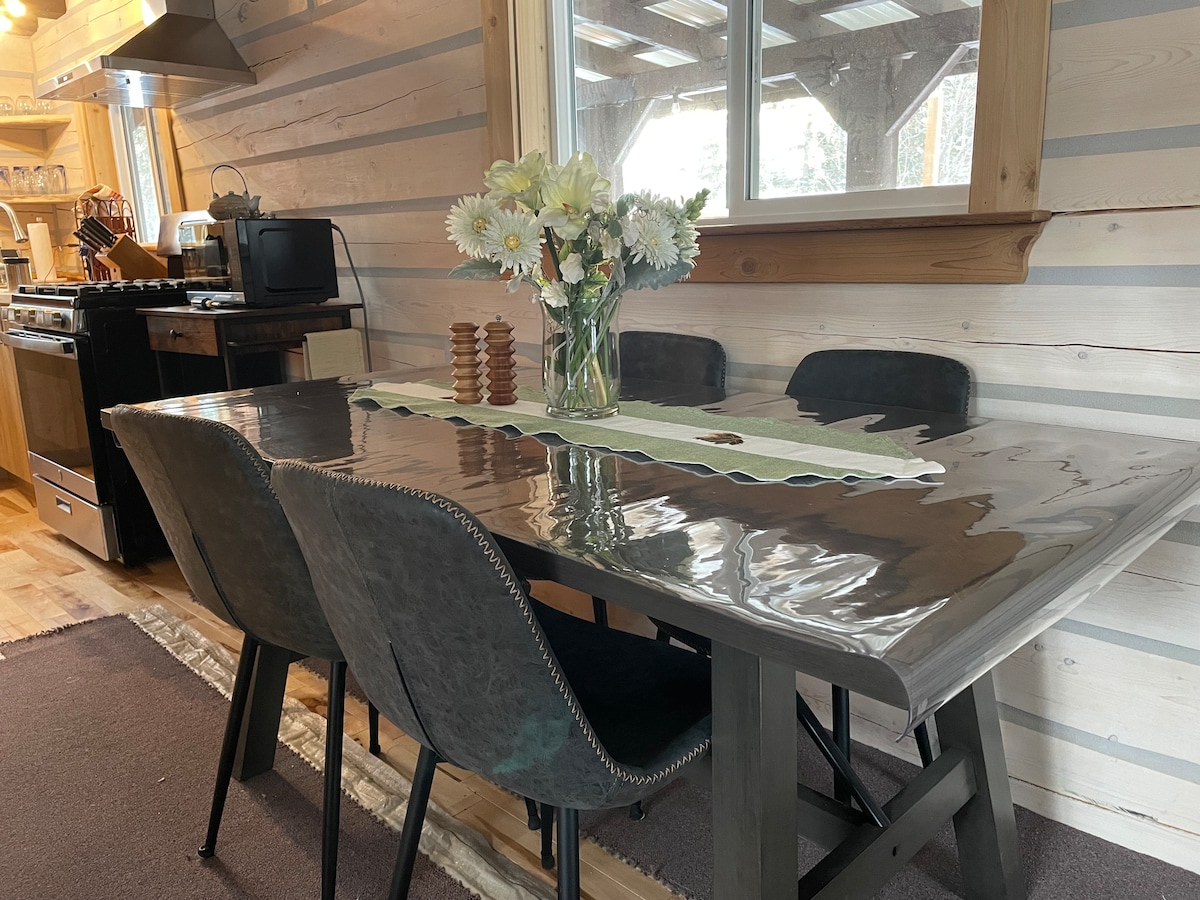 A wooden dining table with a glossy surface is set beneath a window, surrounded by four dark chairs. A vase filled with fresh white flowers adds a touch of nature, while a green table runner and wooden salt and pepper shakers are positioned atop the table.