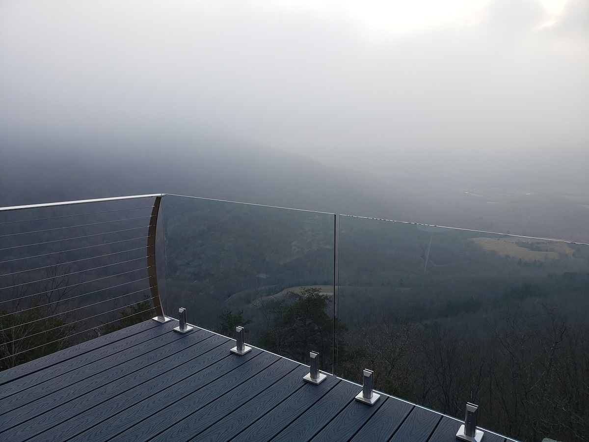 A foggy landscape is showcased from the deck, with the glass railing providing an unobstructed view of the mountains and valley below. The wooden surface of the deck features a textured design, offering a spacious area for relaxation.